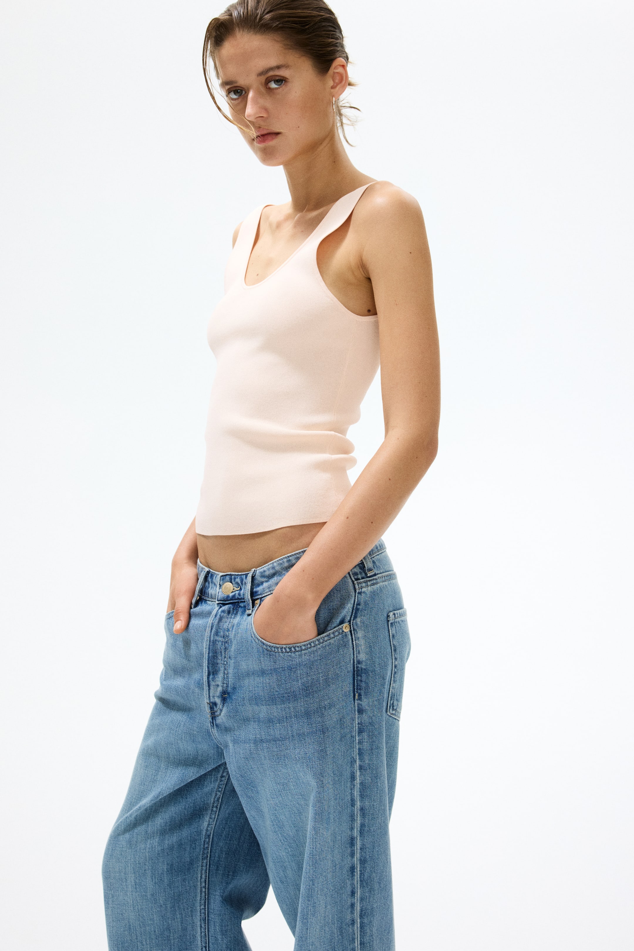 View larger image: A light pink ribbed knit tank top features a scooped V-neck and wide shoulder straps, fitting snugly and ending above the navel. Light blue relaxed fit jeans feature front pockets and a visible button closure, styled with hands in the pockets.