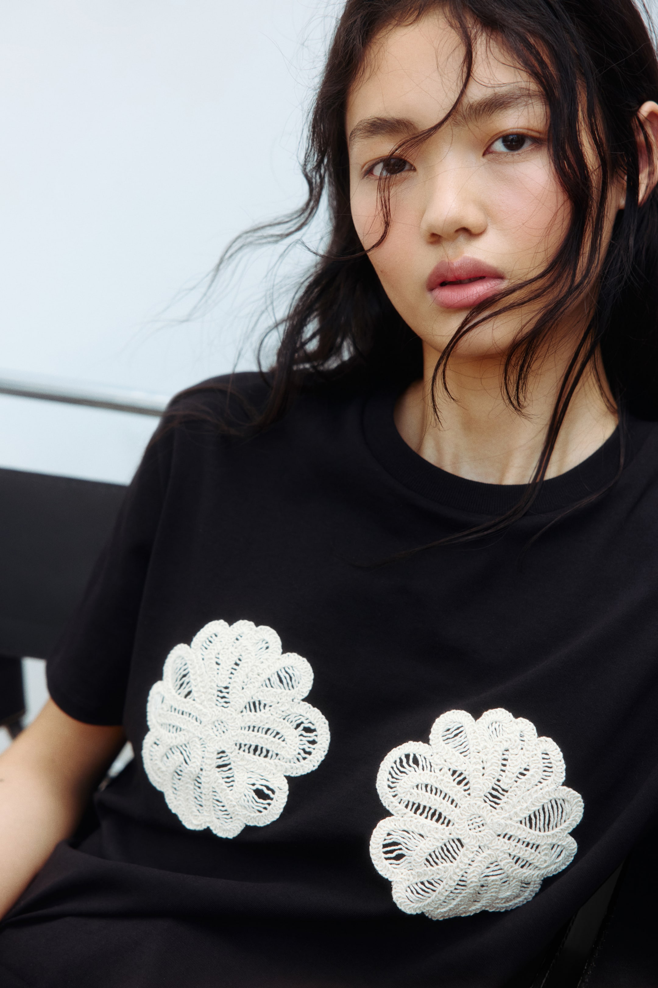 View larger image: This black t-shirt showcases a round neckline and short sleeves, presented from a subtle front-right perspective. The front chest area is adorned with two white, intricately crocheted appliques, reminiscent of layered fan or shell patterns.