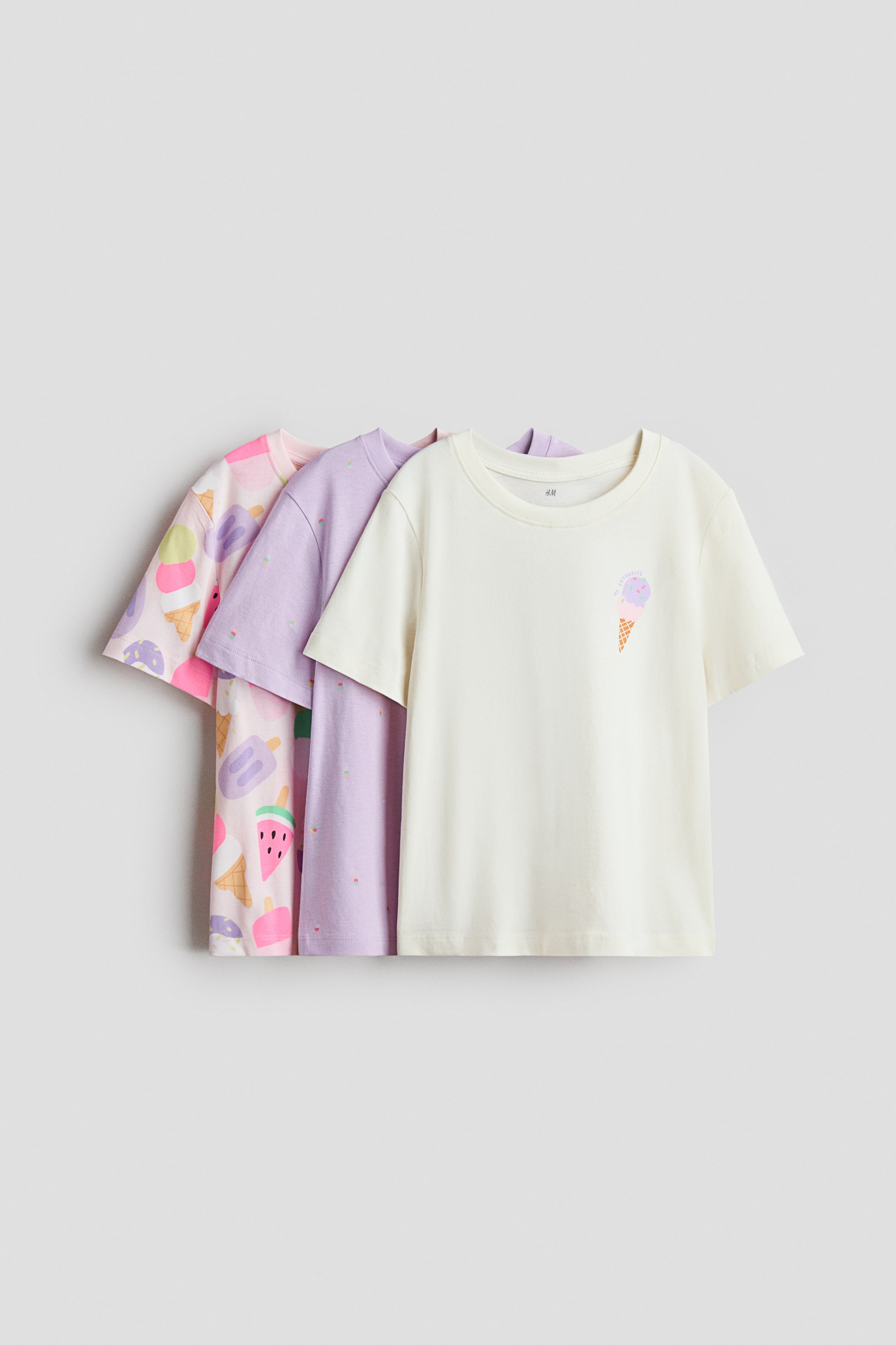 Visualizza immagine più grande: Tre T-shirt in morbido jersey di cotone stampato mostrano la loro vista frontale. La T-shirt color crema più in avanti presenta una grafica con un cono gelato, la T-shirt color lilla chiaro al centro mostra un motivo a pois sparsi, mentre la T-shirt rosa chiaro più indietro presenta una stampa all-over di gelati e frutta. Ogni T-shirt è progettata con un bordo a coste intorno allo scollo e spalle leggermente abbassate.
