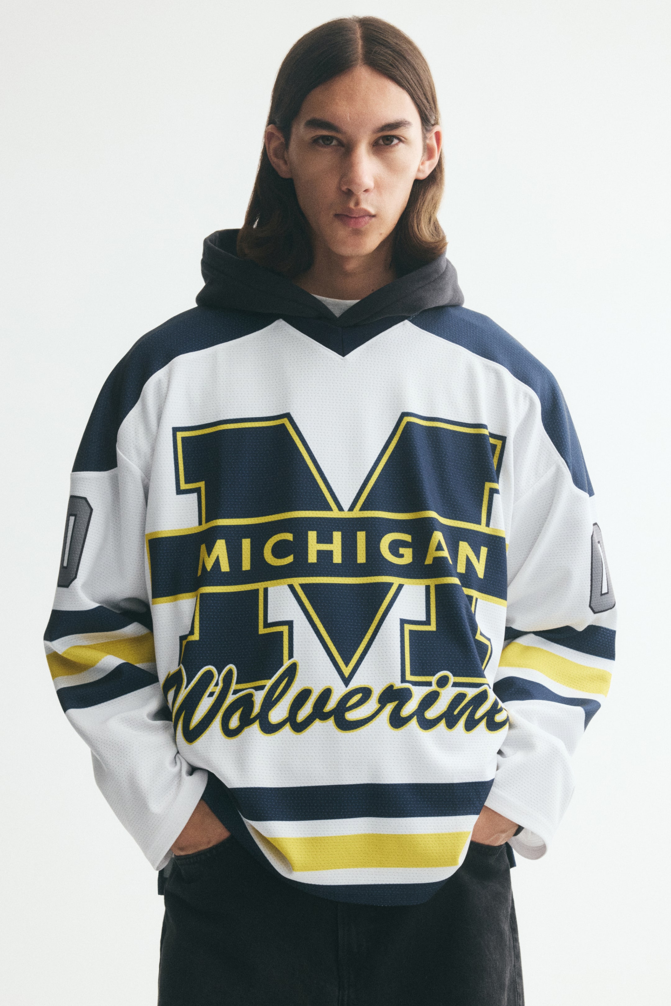 Vezi imaginea mai mare: Un tricou de hochei din plasă cu mâneci lungi, cu un design Michigan Wolverines în alb, bleu marin și galben, este purtat de model peste un hanorac gri închis. Tricoul oversized este stilizat cu umeri căzuți și dungi atletice clasice pe mâneci și la tiv.