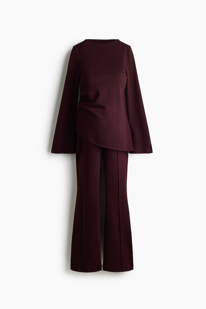 Un conjunto de jersey de dos piezas en color burdeos se muestra de frente, compuesto por un top de manga larga y un pantalón a juego. El top presenta cuello alto tipo mock neck, mangas largas acampanadas, drapeado lateral en la cintura y un bajo curvo asimétrico. El pantalón presenta tiro alto y perneras acampanadas con pliegues centrales cosidos que recorren la parte delantera. - 1