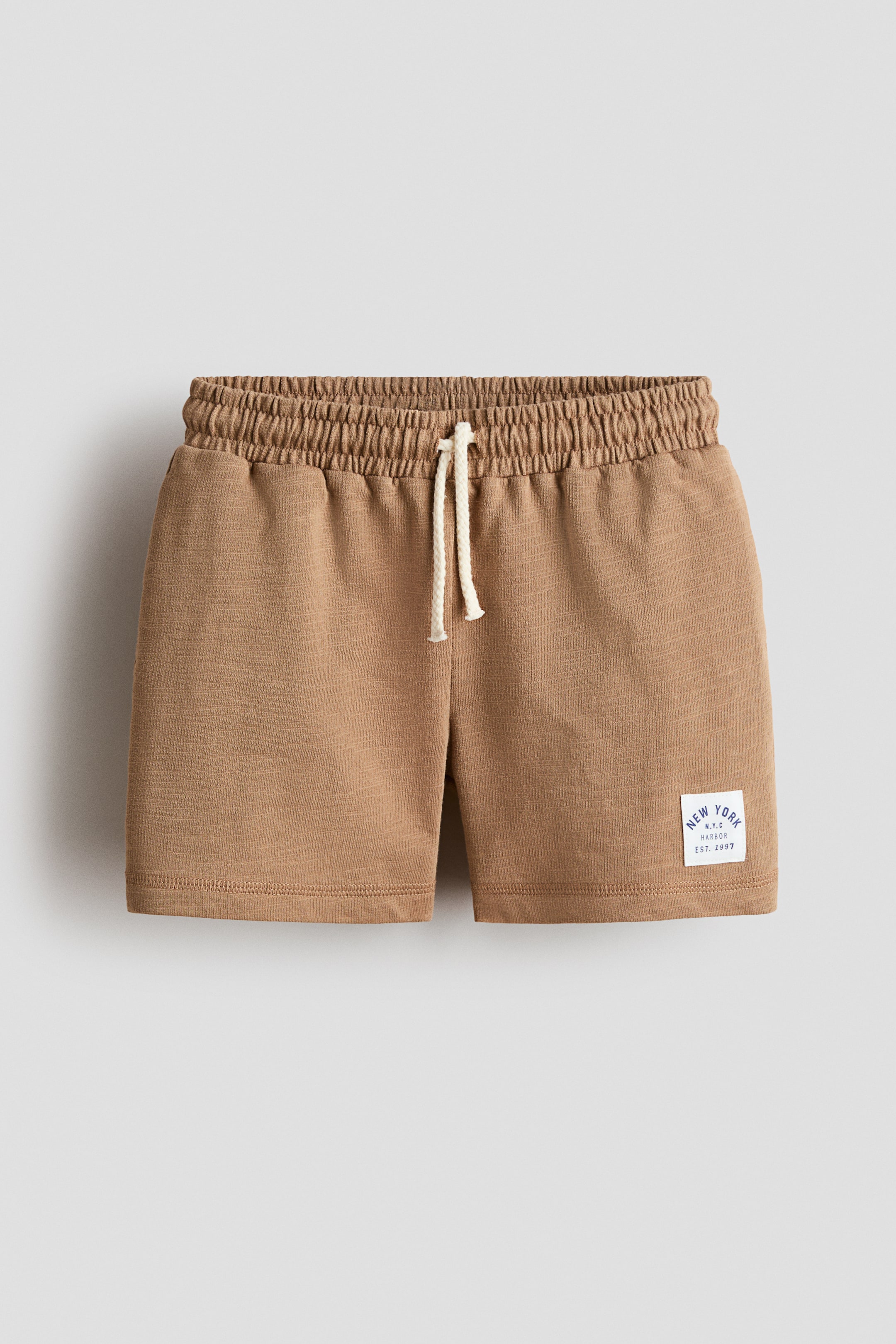View larger image: Front view of brown shorts showcasing an elasticated waistband with a light-coloured drawstring tied in a bow. A white rectangular patch displaying "NEW YORK N.Y.C. HARBOUR EST. 1997" text is visible on the lower right leg.