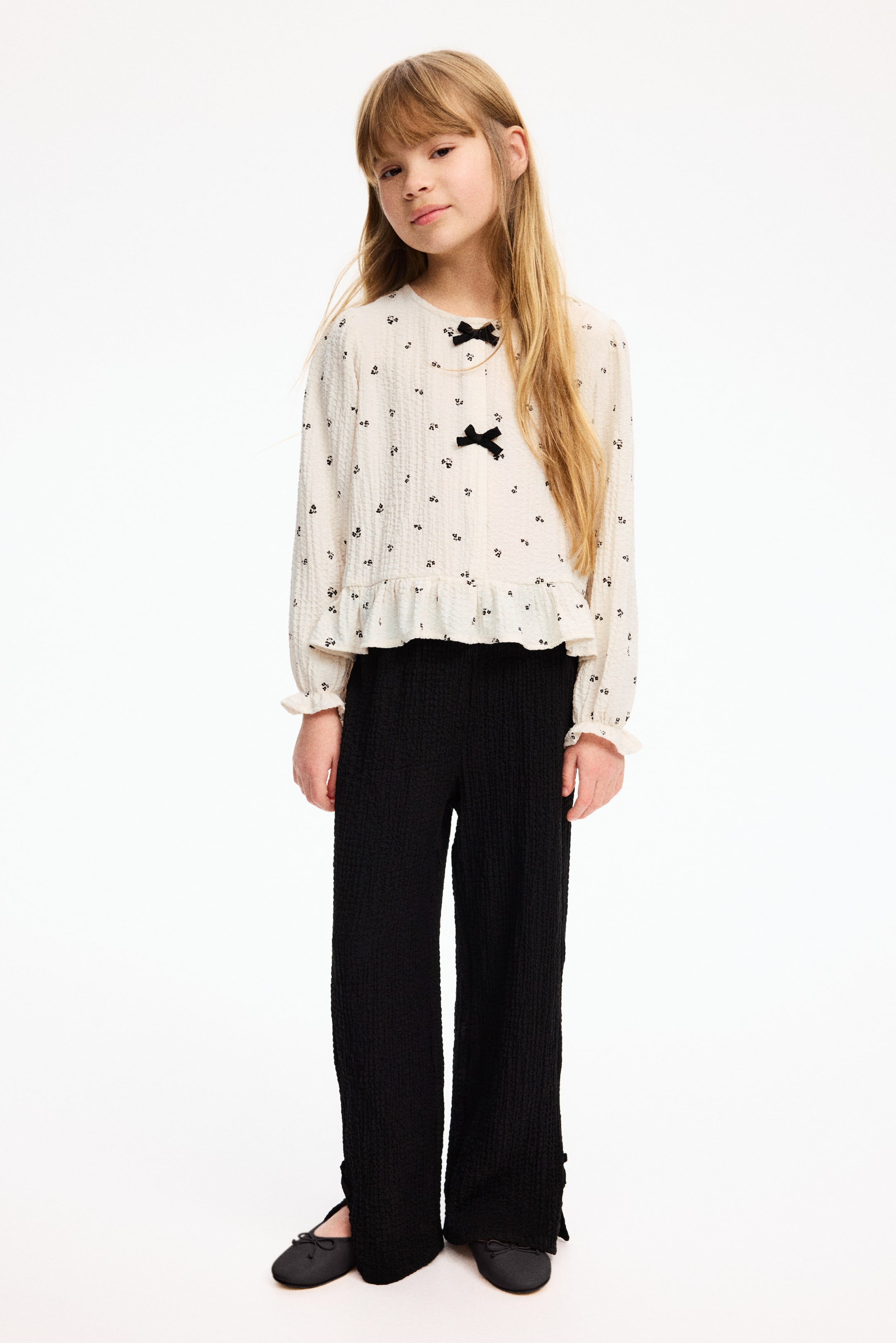 View larger image: A cream-textured long-sleeve top with small black floral prints, a ruffled peplum hem and ruffled cuffs is paired with black wide-leg textured pants. The top features a round neckline and two decorative black bows at the front, while the pants have split hems with button details at the ankles.