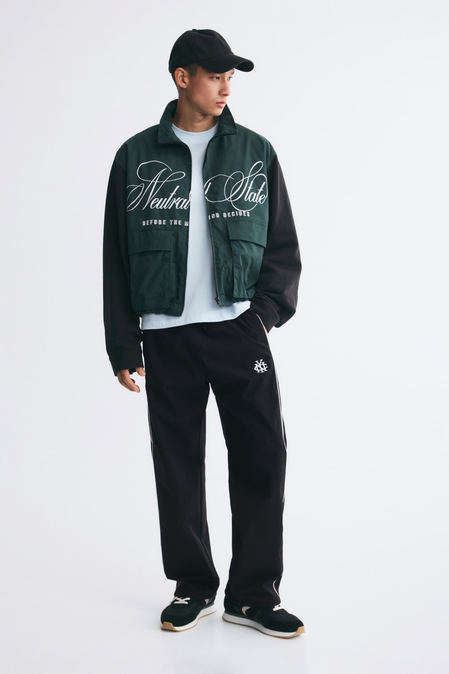 Black track pants featuring a relaxed fit, a small white embroidered logo on the left thigh, and thin white piping down the sides are being worn by the model. The pants are styled with a green and black graphic jacket, a light blue t-shirt, a black baseball cap, and sneakers. - 1