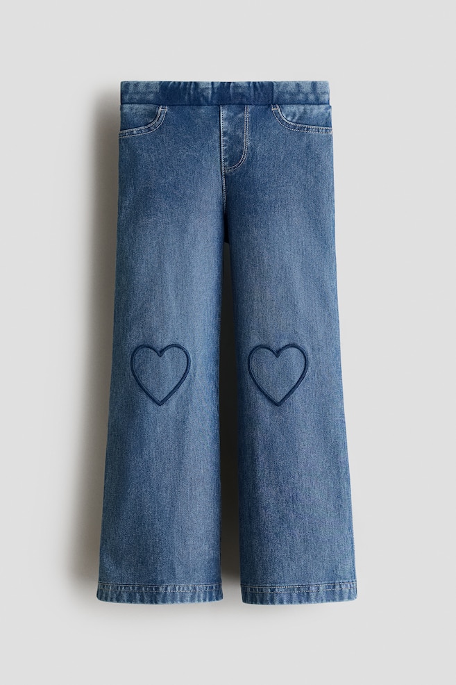 A pair of blue denim-look jersey jeggings is shown in a front-facing view, featuring an elasticated waistband, a mock fly, and decorative front pockets. The relaxed-cut legs display embroidered navy blue heart outlines on each knee. - 1