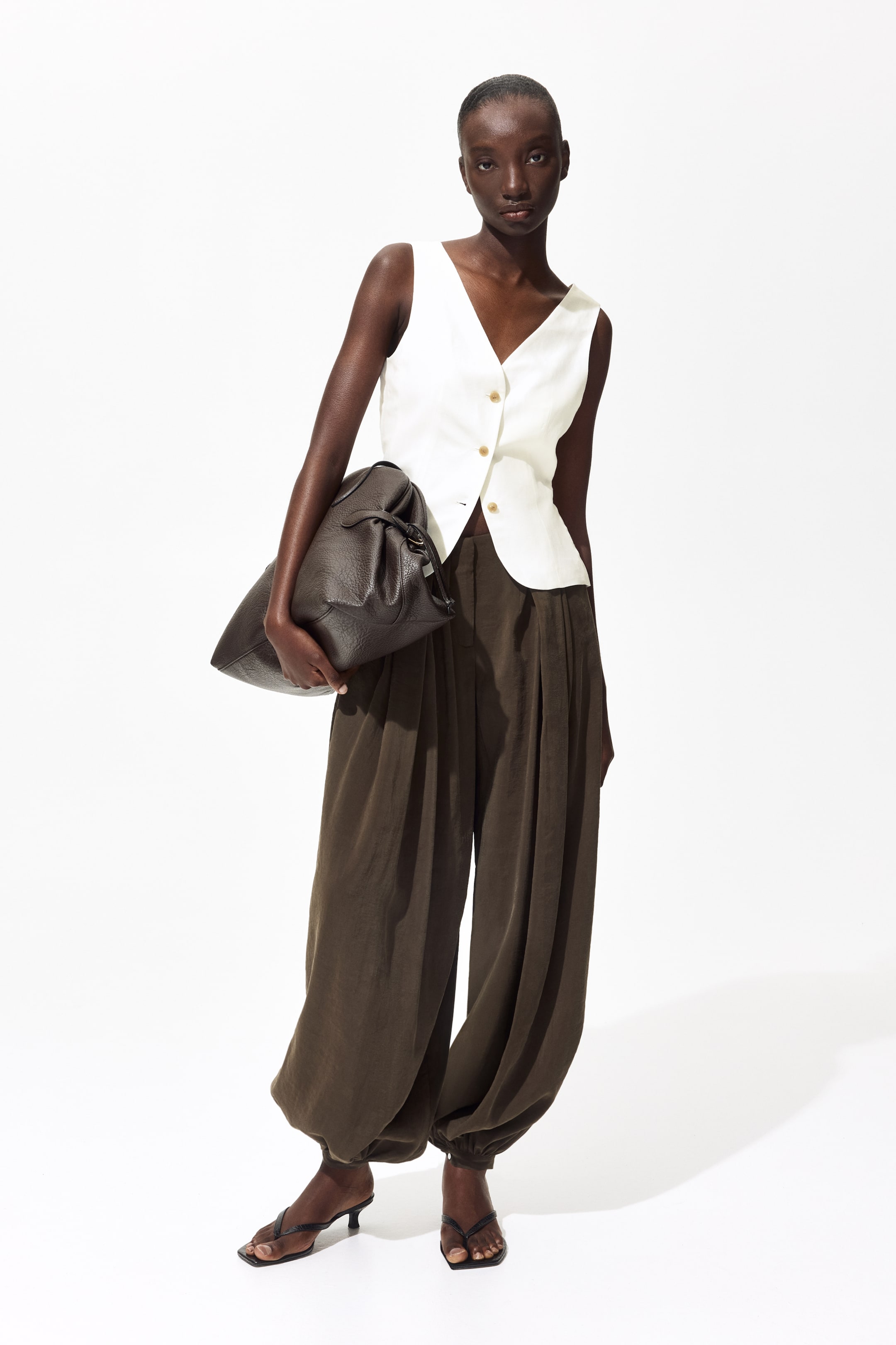 View larger image: The model is wearing a white tailored vest crafted from a viscose and linen blend. It boasts a V-neckline and gold-tone buttons along the front, some of which are partially fastened. The vest is styled with wide-leg pants.
