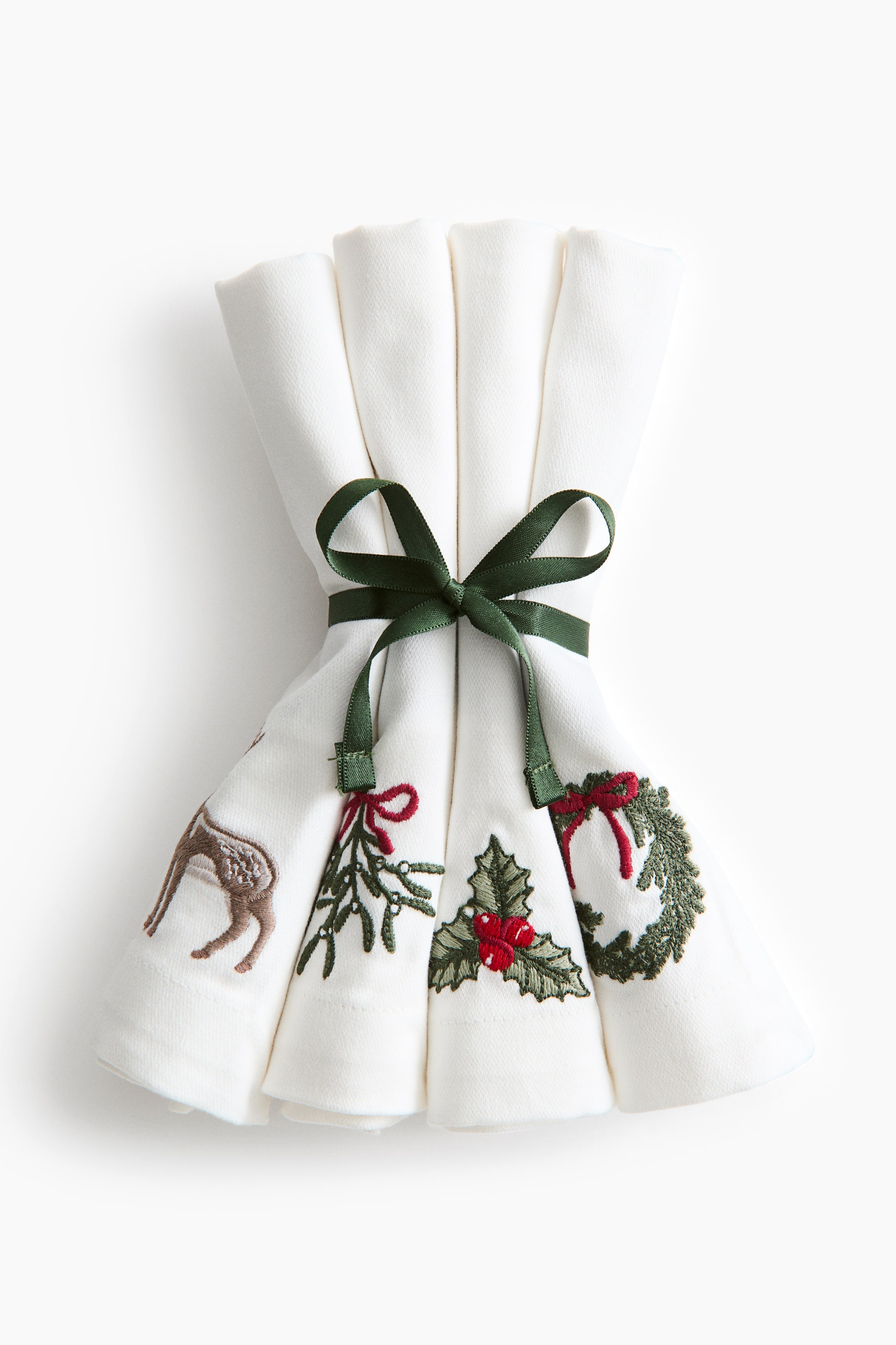 View larger image: A set of four white fabric napkins is arranged upright, bundled together and tied in the middle with a dark green ribbon bow. Facing front, each folded napkin displays a unique embroidered design along the bottom edge, including a brown deer, a mistletoe sprig with a red bow, holly leaves with red berries, and a green wreath with a red bow.