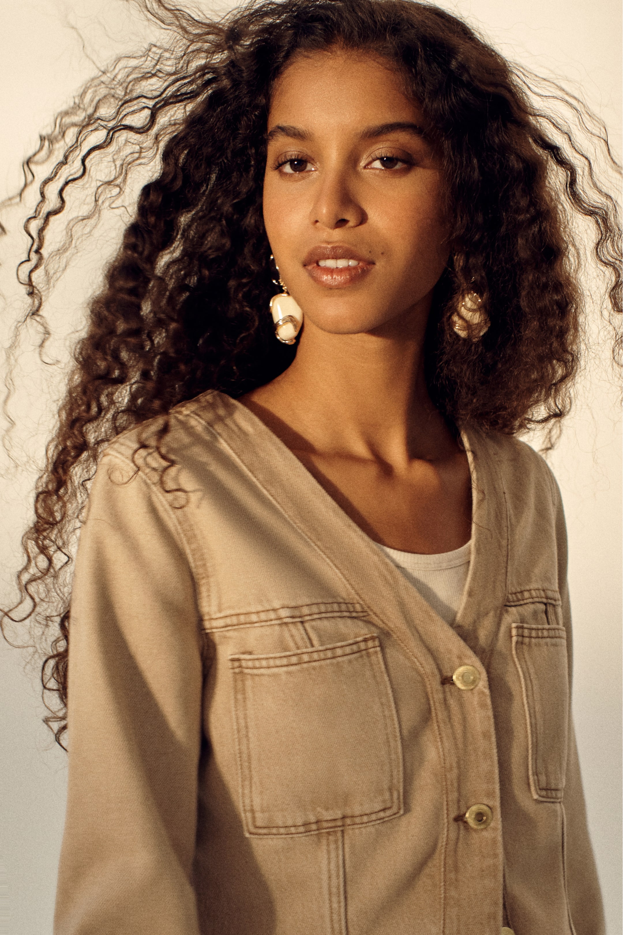 Ampliar la imagen: Una chaqueta beige con cuello en V que presenta un cierre frontal de botones con botones de color claro, dos grandes bolsillos de parche y costuras prominentes en todo su diseño. Se lleva sobre una blusa blanca.