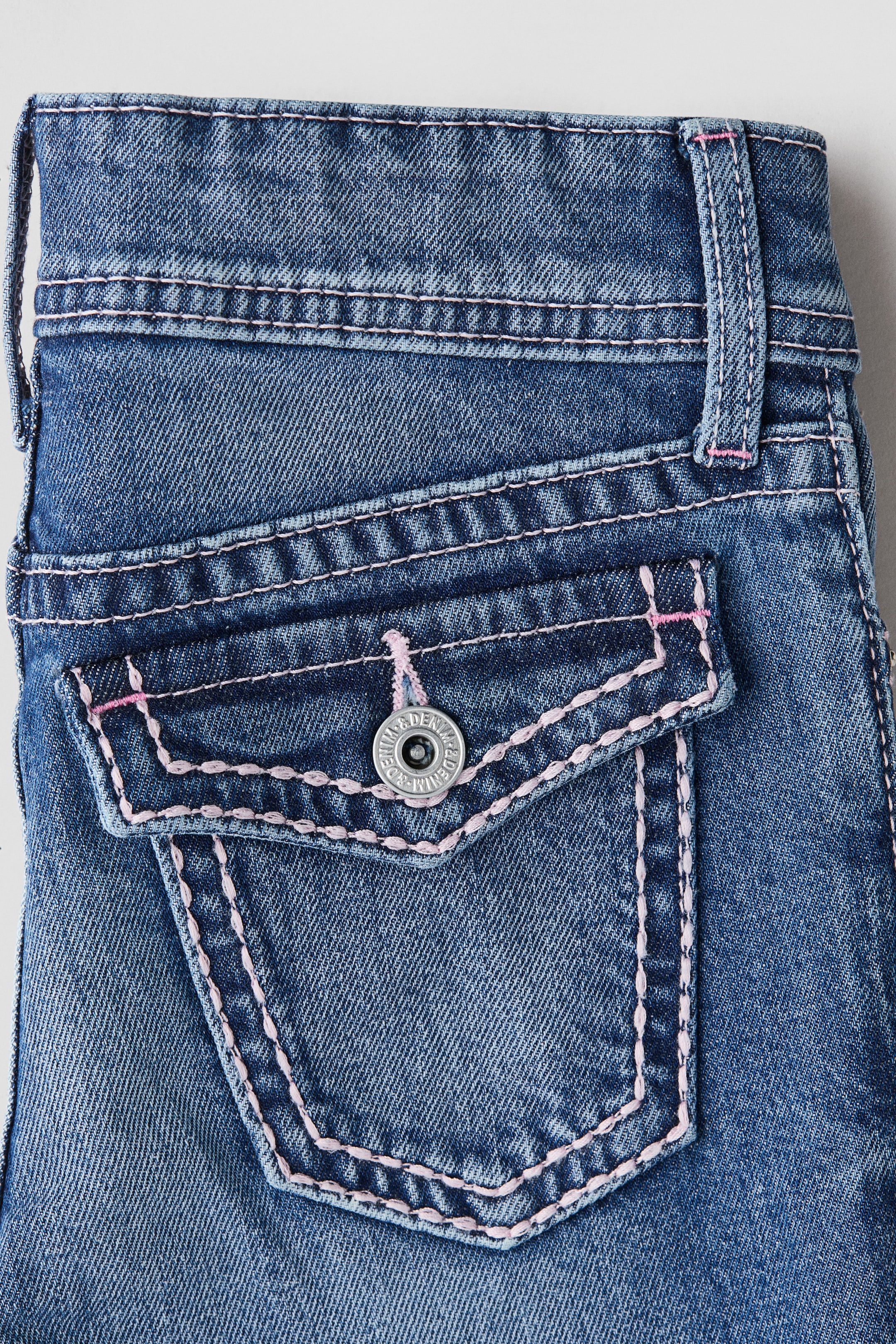 View larger image: A close-up view of blue denim jeans is showcasing a back pocket with a buttoned flap. The pocket and flap are being outlined with decorative pink contrast stitching, and a silver-toned button is securing the flap.