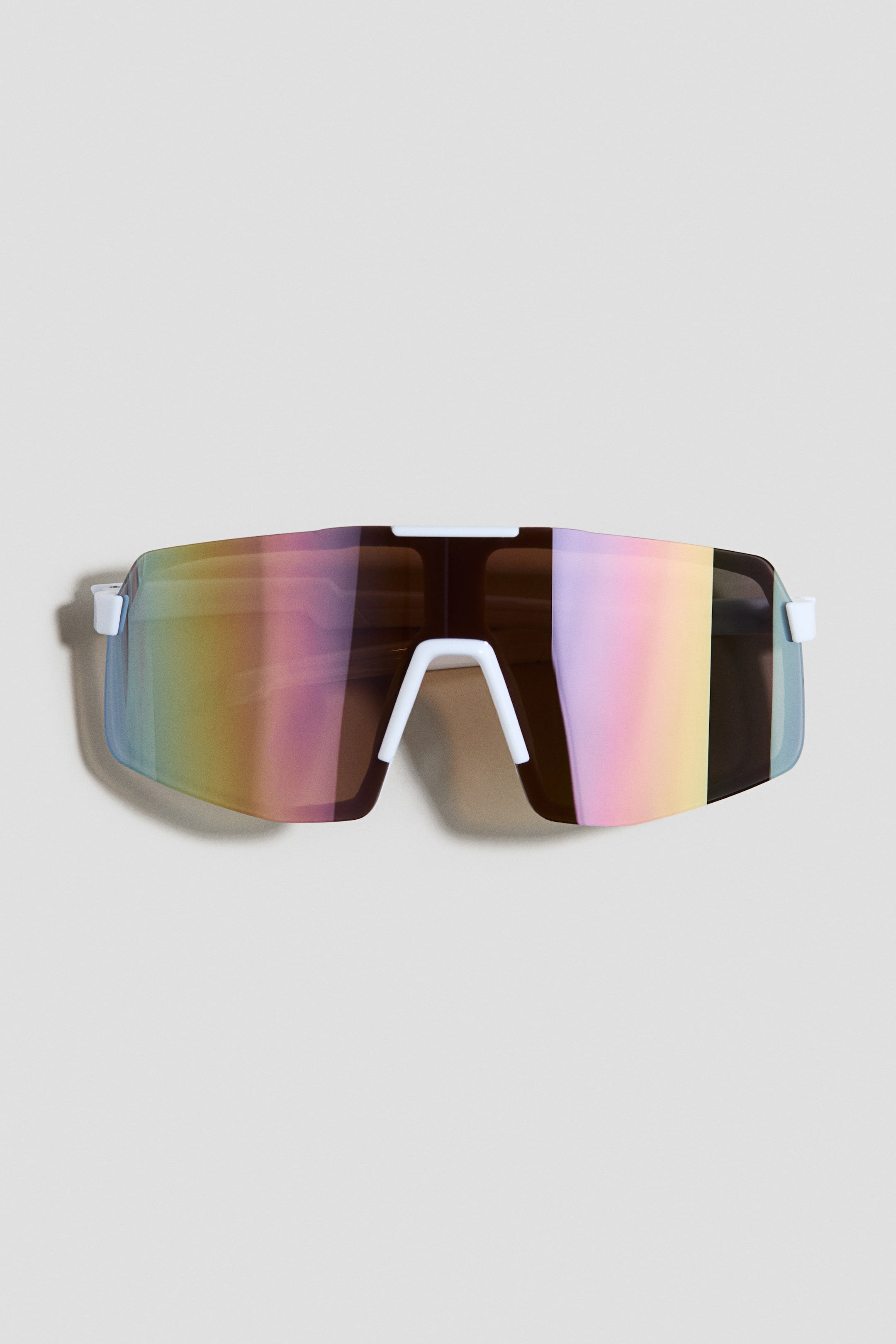 View larger image: Sunglasses are displayed in a FRONT view, featuring a large, single shield lens that is tinted and mirrored, showcasing an iridescent rainbow effect with hues of pink, yellow, blue, and green. A white plastic frame forms the top edge and a prominent nose bridge.