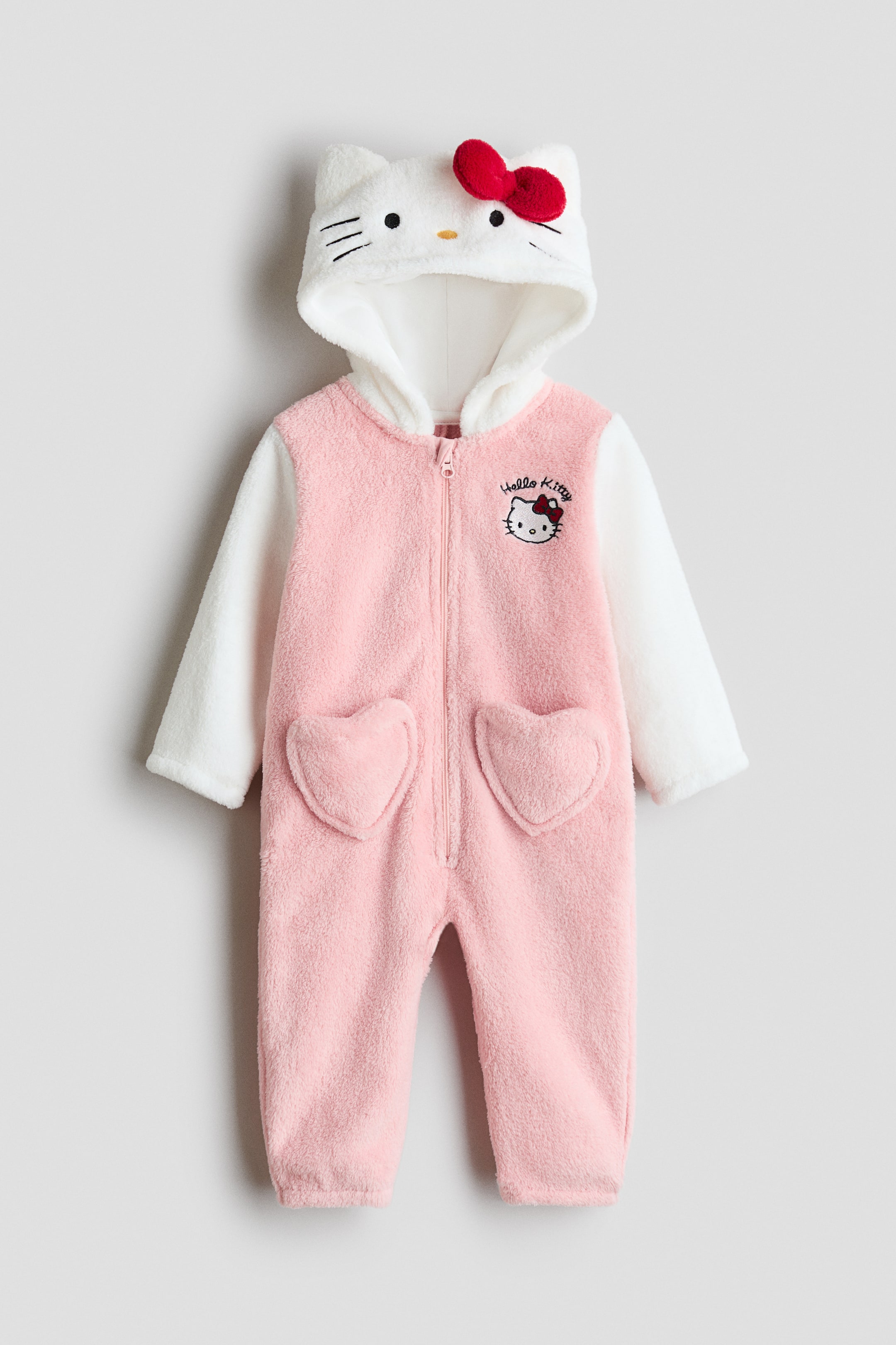 View larger image: The Hello Kitty baby onesie is showcasing a full-length, front-facing view. It boasts a pink plush body with white long sleeves and a white hood. The hood is crafted as Hello Kitty's face, complete with embroidered eyes, whiskers, and a red bow, while two heart-shaped pockets and a Hello Kitty logo embellish the front.