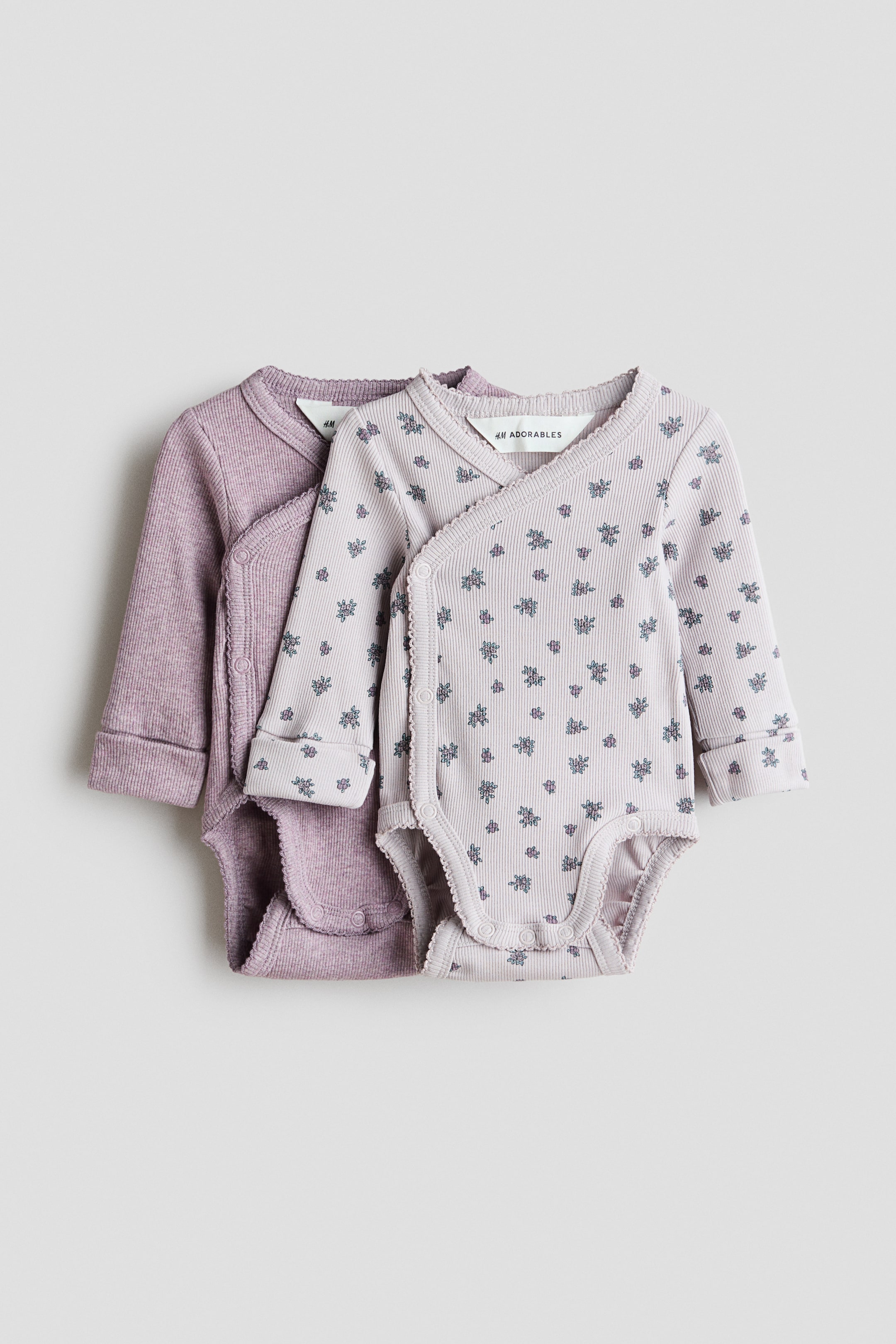 查看大圖: 兩件 H&M Adorables 嬰兒連身衣稍微重疊地平放，均展示羅紋針織面料和長袖設計。前面的連身衣展示淺薰衣草色花卉圖案，而後面的連身衣則呈現純薰衣草色。兩件連身衣均採用交叉前襟設計，側面有按鈕扣，袖口摺疊，底部有雙排按鈕扣。