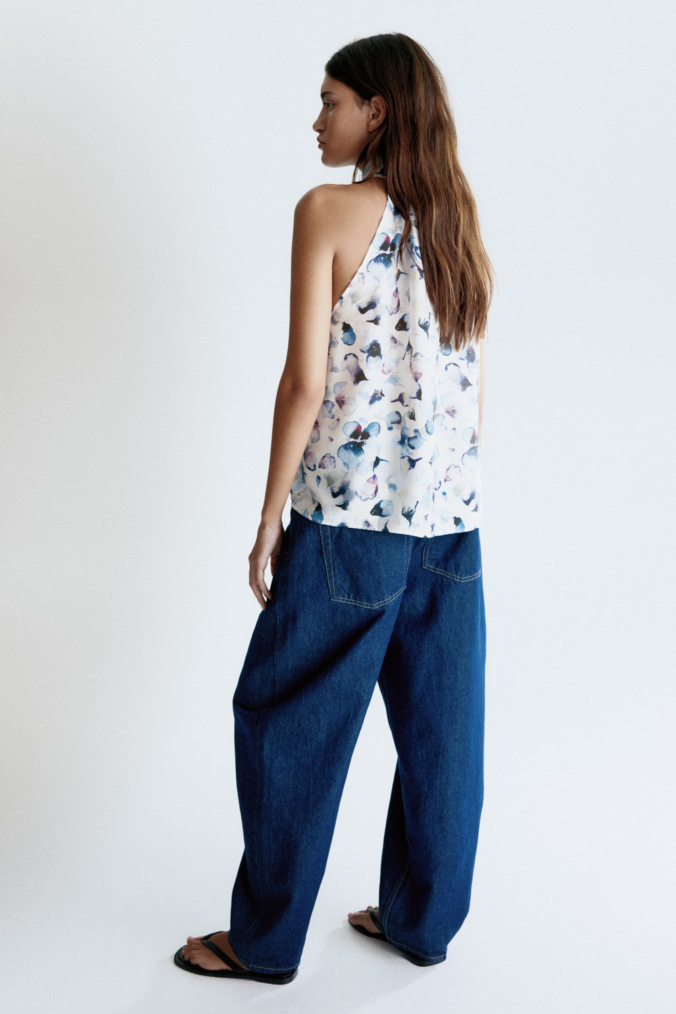 View larger image: A sleeveless halter-neck top is being shown from a back and side angle. It is featuring an abstract floral pattern in blue and purple on a light background and is flowing loosely over dark blue, wide-leg denim pants, which are showcasing visible stitching.