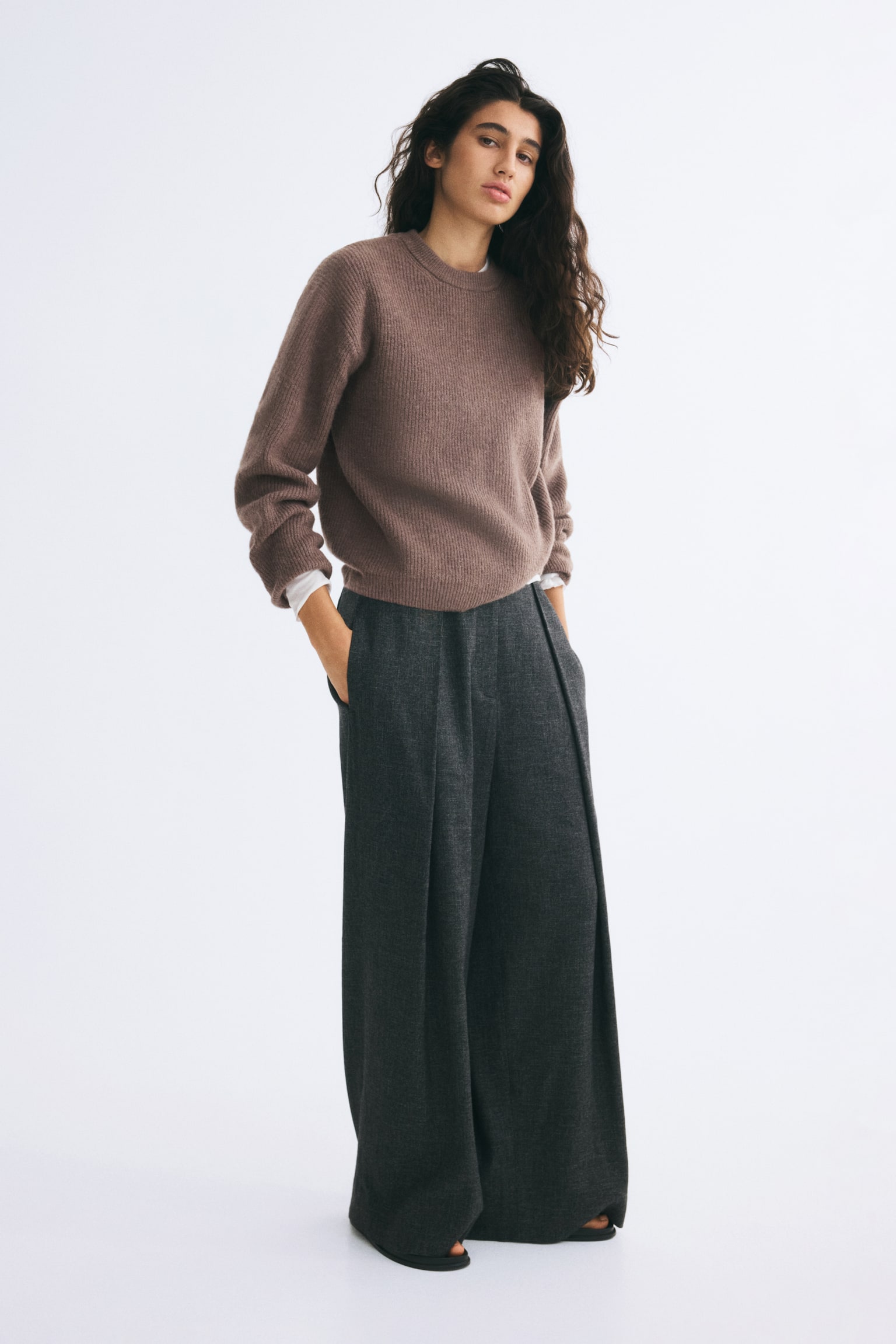 Un pull en maille douce marron, avec un col rond cÃ´telÃ©, des Ã©paules tombantes et des poignets cÃ´telÃ©s, est portÃ© par le mannequin. Le pull est superposÃ© Ã  un haut blanc et associÃ© Ã  un pantalon large gris foncÃ©. - 1