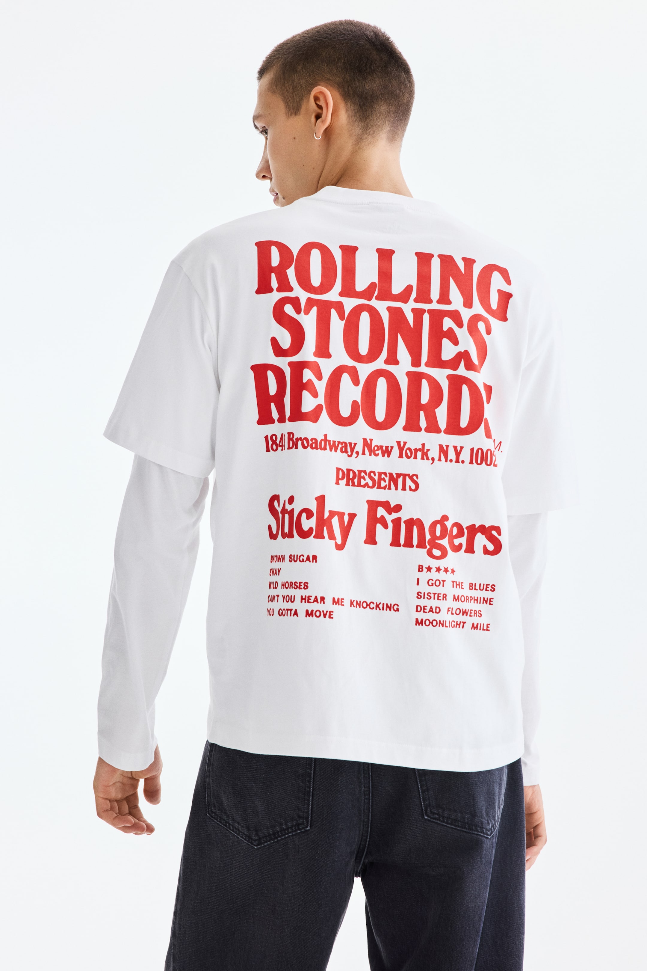 Grotere afbeelding bekijken: Een wit T-shirt met korte mouwen wordt over een wit shirt met lange mouwen gedragen, gezien vanaf de achterkant. Het T-shirt heeft grote rode tekst op de achterkant die luidt: 'ROLLING STONES RECORD PRESENTS Sticky Fingers' met een adres en een lijst van songtitels.