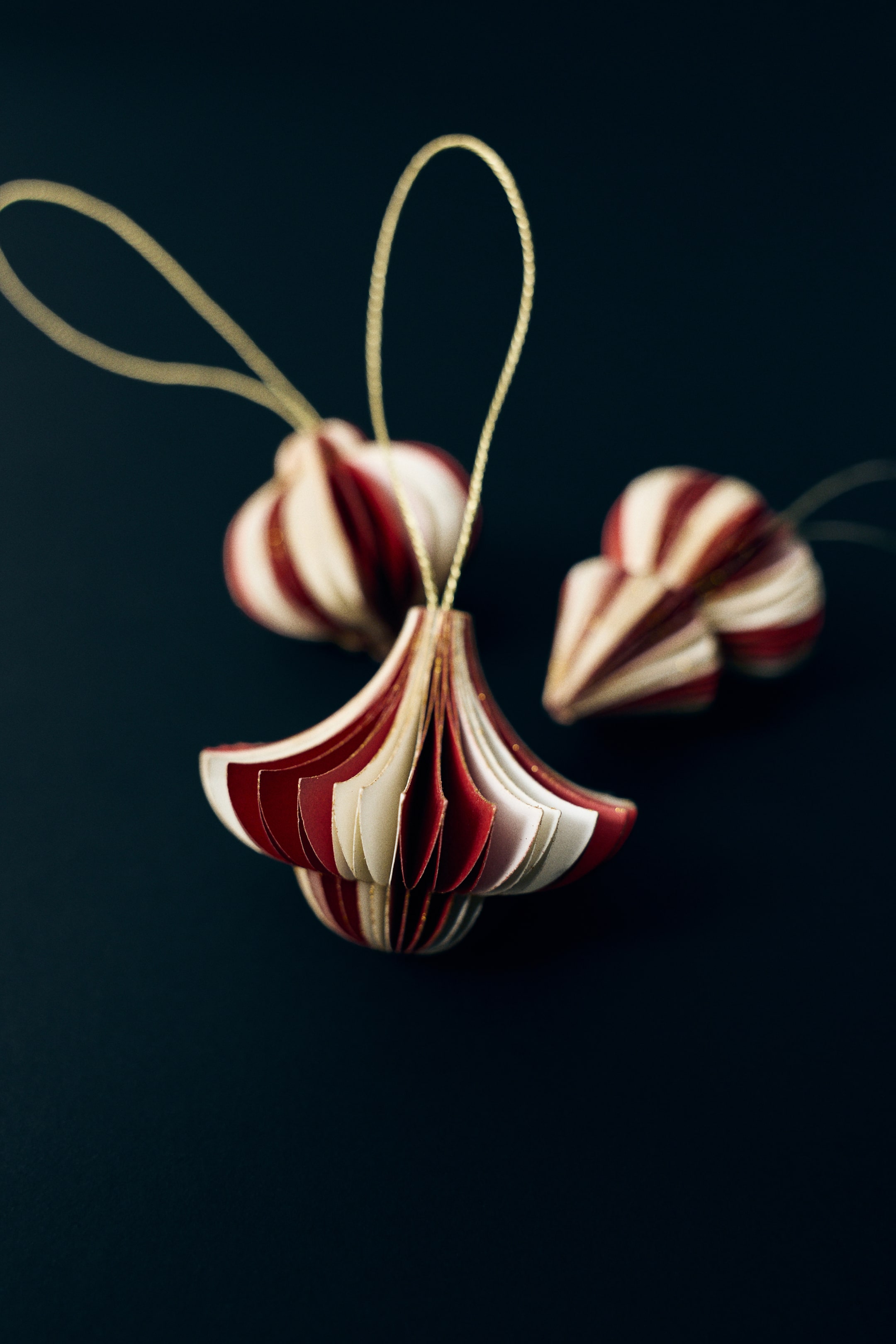 Agrandir l'image: Des ornements de Noël en papier présentent un design tridimensionnel à plis accordéon, avec des couches de papier rouge et crème alternées. L’ornement principal est orienté vers l’avant, mettant en valeur sa forme en bulbe, ses bords recouverts de paillettes dorées et une fine boucle de ficelle dorée fixée en haut pour la suspension.