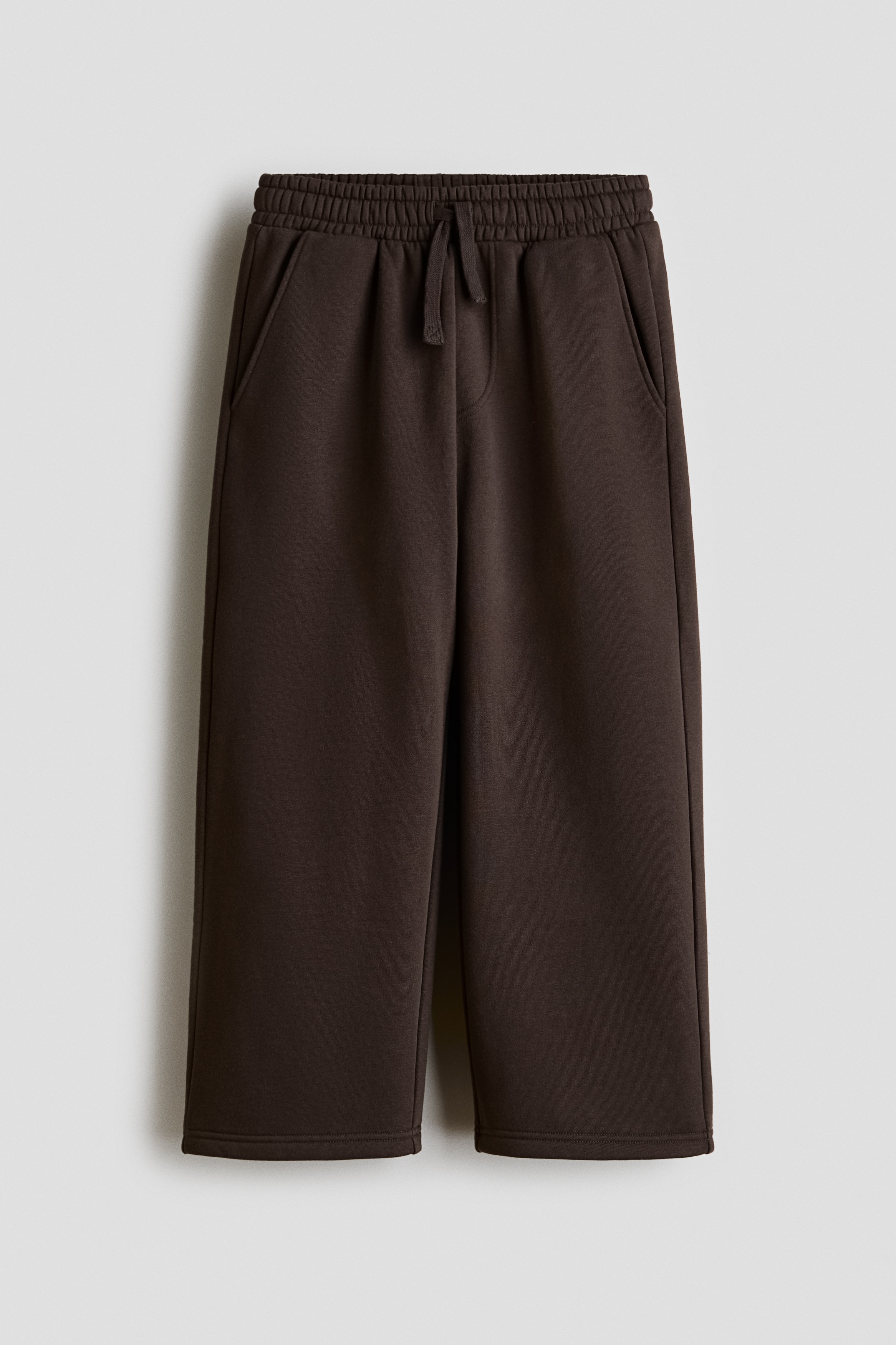 Dark brown track pants are shown from a front-facing angle, featuring a relaxed, wide-leg silhouette and an elasticated waistband with a drawstring. The pants are draping loosely over sneakers, styled alongside an open light grey zip-up hoodie and a white t-shirt.