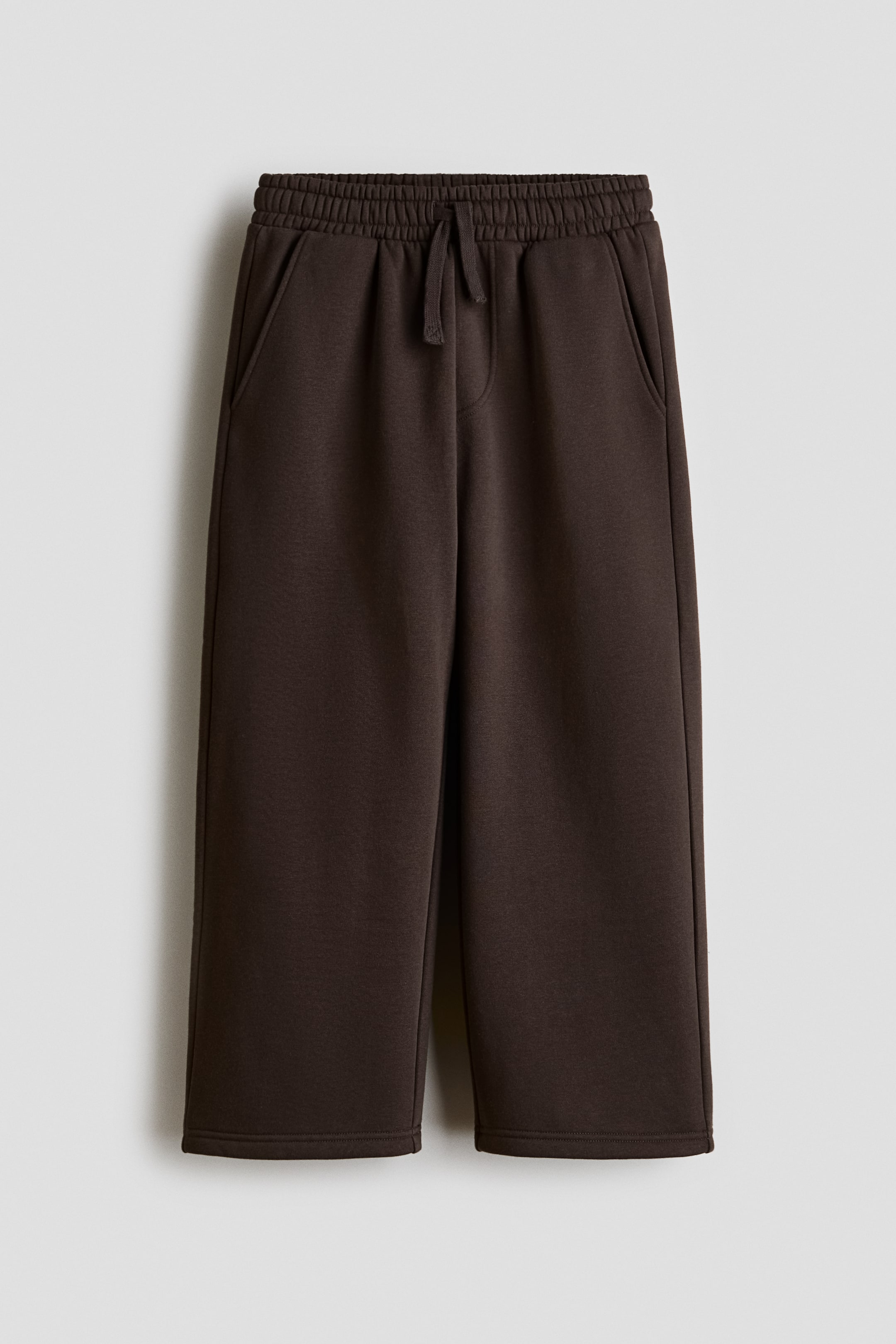 Grotere afbeelding bekijken: Donkerbruine joggers worden van voren getoond, met een elastische tailleband met trekkoord en gestrikte koordsluiting. De joggers hebben schuine steekzakken aan de zijkant en wijde pijpen.