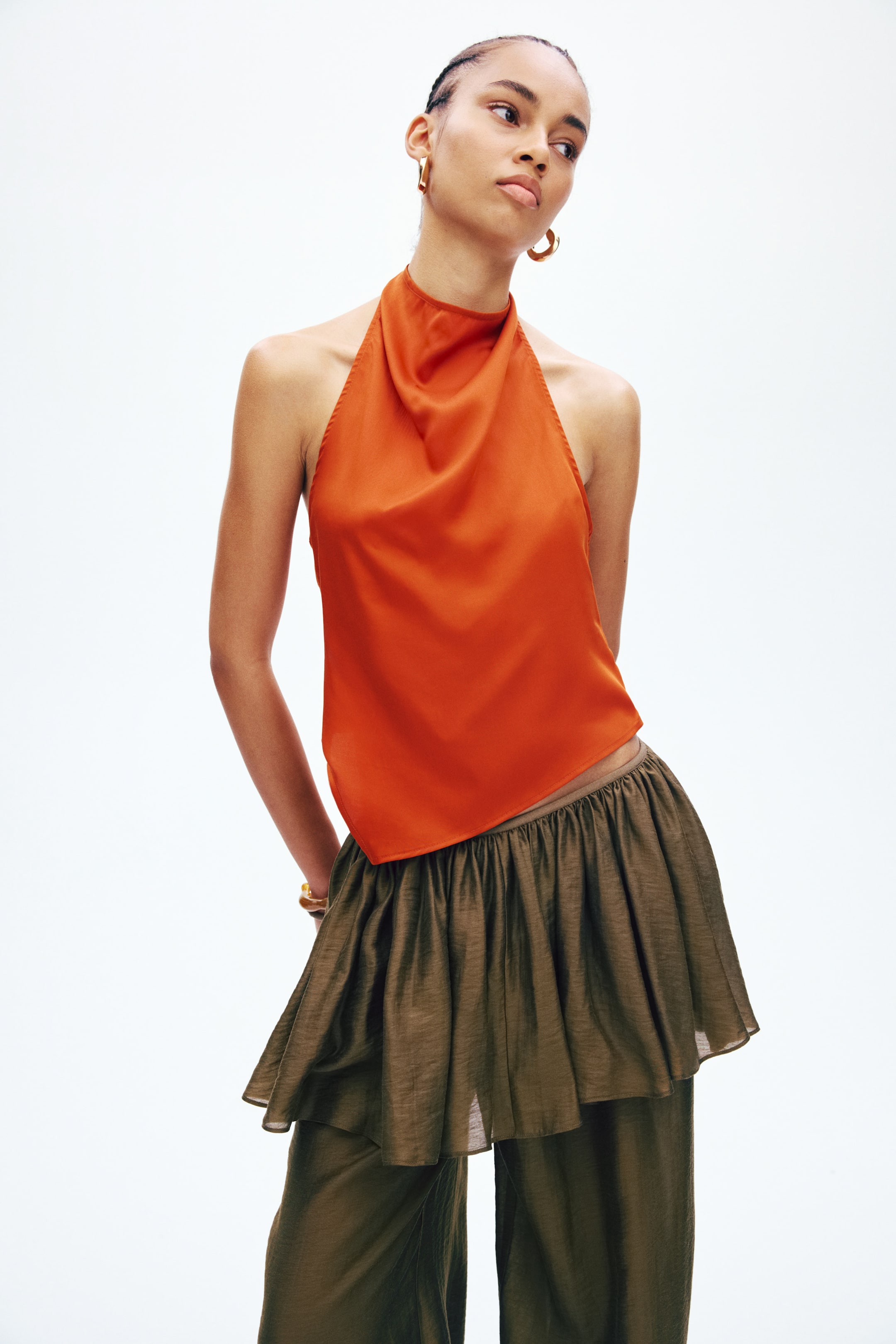 View larger image: An orange satin halter top showcases a draped cowl neck and an asymmetrical hemline, layered over olive green wide-leg trousers that feature a gathered peplum overlay at the waist, with the entire ensemble presented from a slight front-right angle.