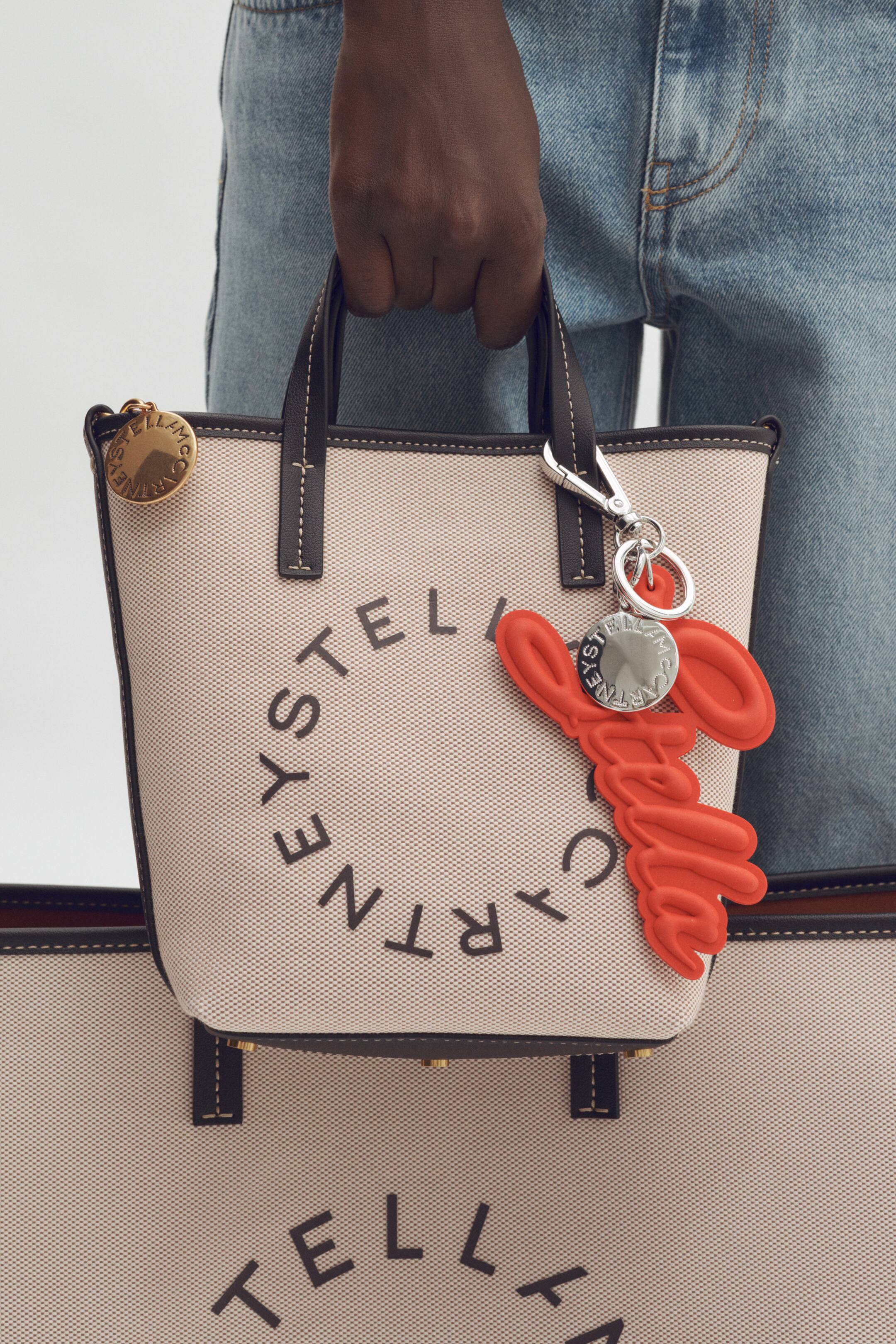 Agrandir l'image: Un sac fourre-tout en toile beige est tenu, doté de poignées marron foncé avec des coutures contrastées blanches. Le devant affiche "STELLA MCCARTNEY" en texte noir disposé en design circulaire, tandis qu'un charme circulaire doré pend de la base d'une poignée et un charme rouge sculpté "Stella McCartney" avec un disque argenté est attaché via un fermoir mousqueton à l'autre.