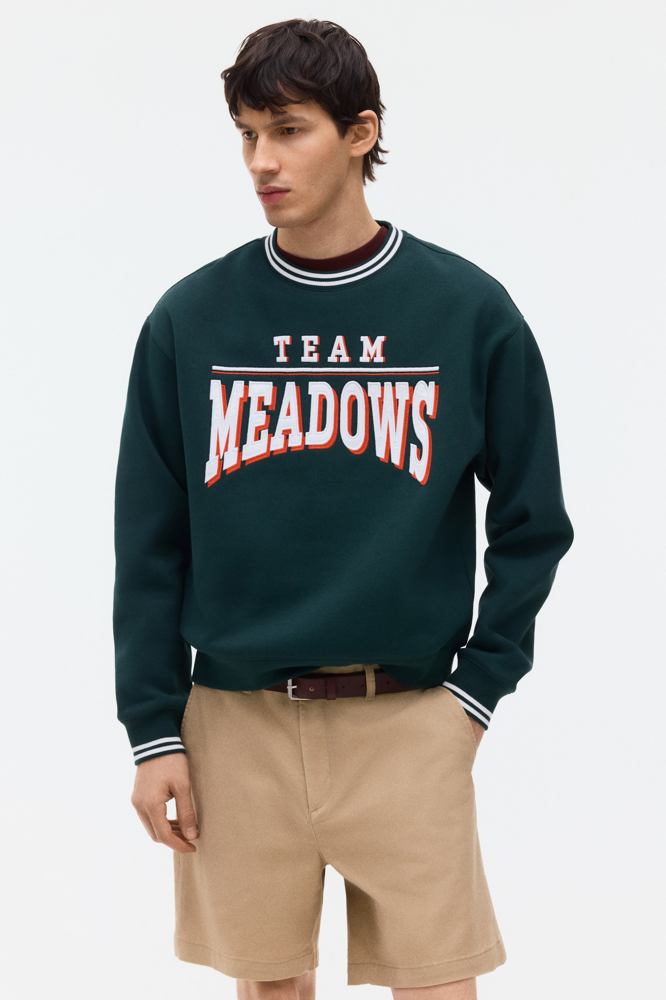 View larger image: A dark green sweatshirt features a ribbed crew neck and ribbed cuffs, each accented with two white stripes. The front showcases "TEAM MEADOWS" in white and orange lettering, with a dark red layer peeking from beneath the collar.