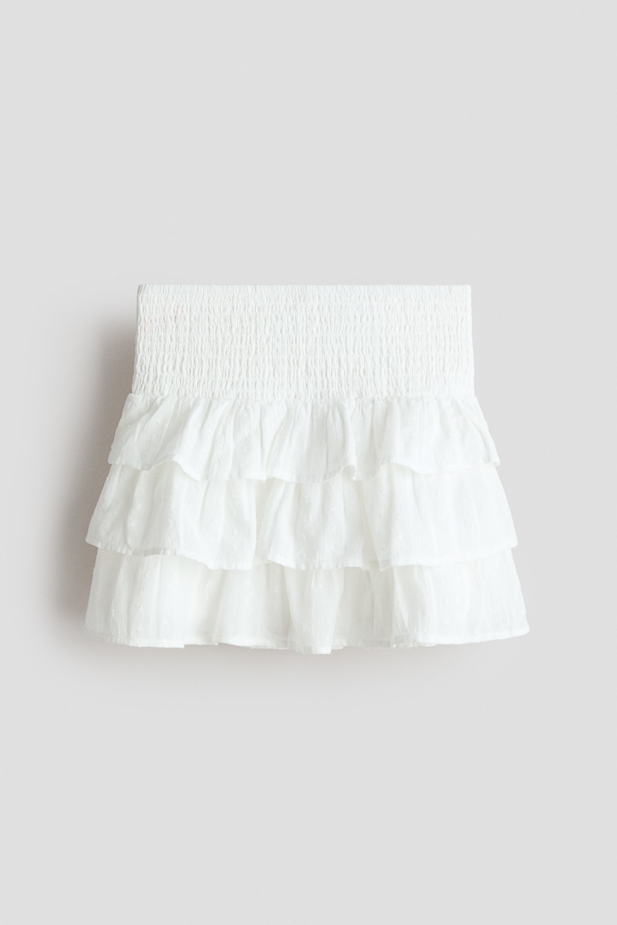 Visualizza immagine più grande: Una gonna a balze bianca presenta un cinturino arricciato in vita e tre strati arricciati. Il tessuto bianco mostra un delicato motivo a pois svizzeri.