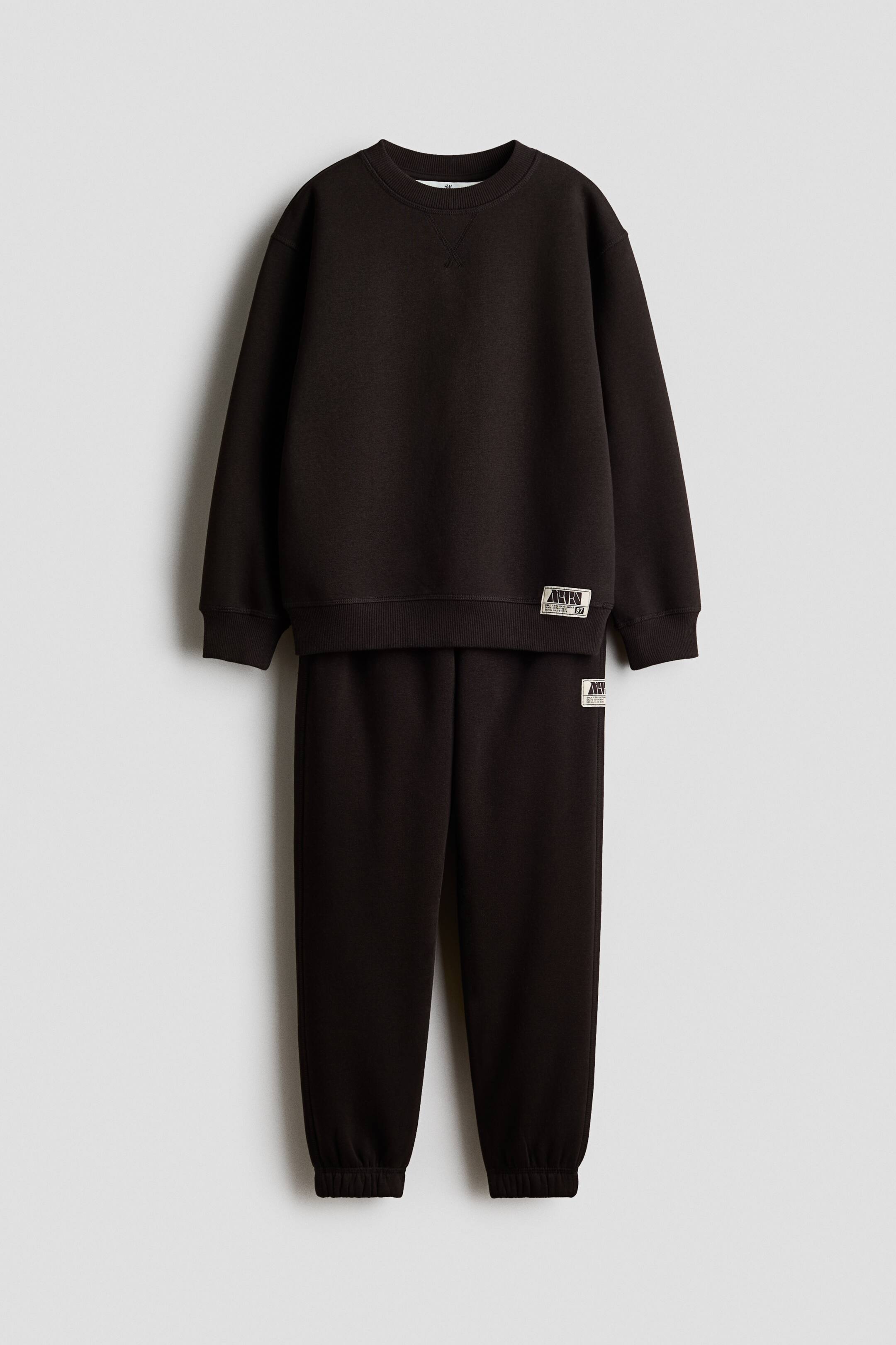 View larger image: The 2-piece brushed-inside sweatshirt set features a dark brown crew-neck sweatshirt with a V-stitch detail below the neckline, ribbed cuffs, and a ribbed hem, along with a decorative patch on the lower left front. The coordinating dark brown sweatpants feature ribbed ankles and a matching decorative patch on the upper left thigh, all shown from the front.