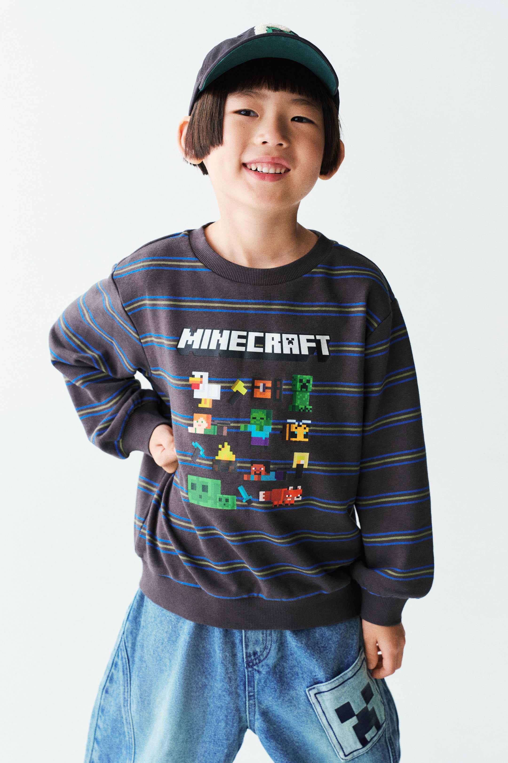 拡大画像を表示: モデルは、プリントされたMinecraftグラフィックがあしらわれたダークグレーのストライプ柄長袖セーターを着用しています。セーターは、リブ仕様のラウンドネックライン、ドロップショルダー、袖口と裾のリブトリムが特徴で、ブルーデニムジーンズとキャップを合わせてスタイリングされています。