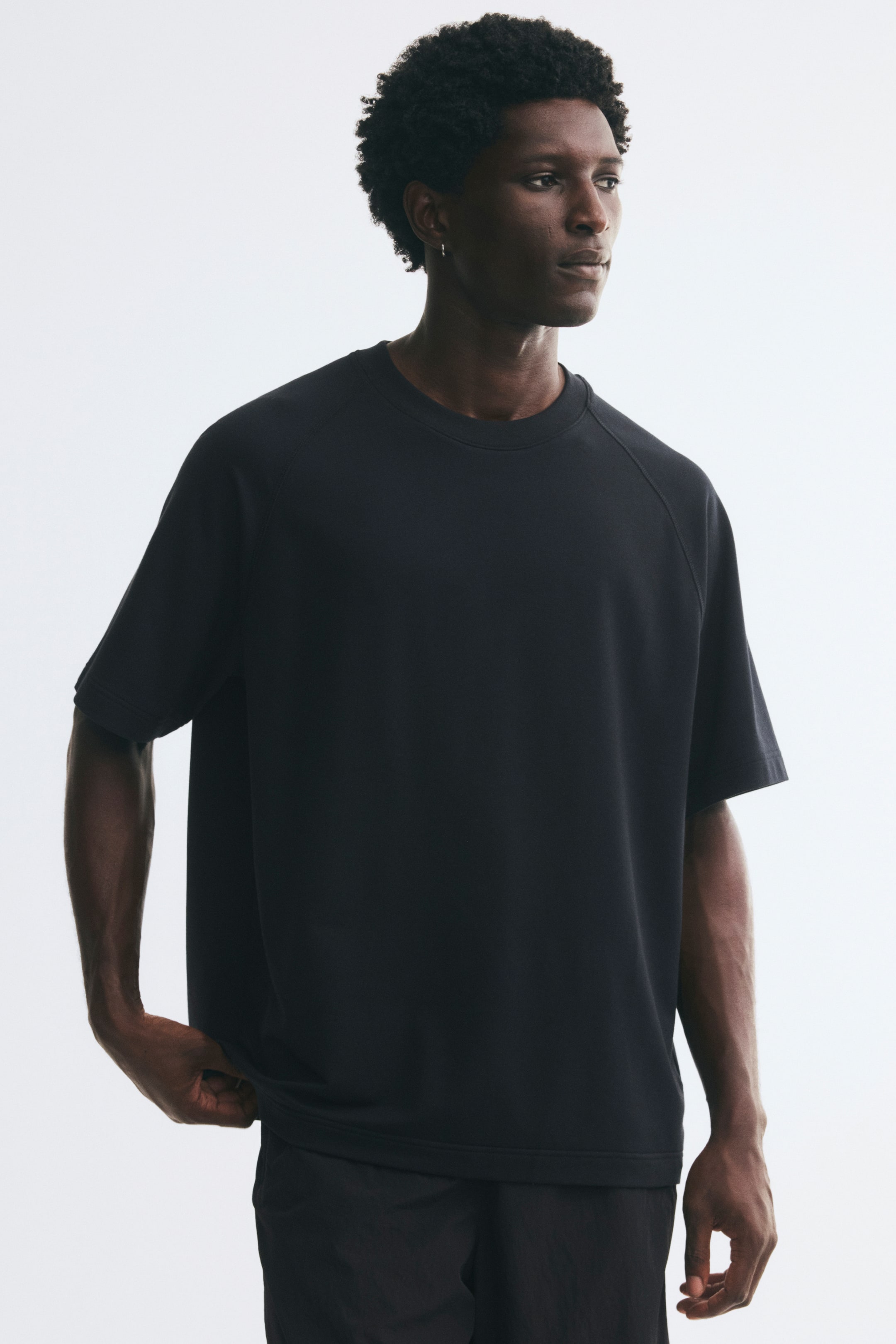 Voir une image plus grande: Un t-shirt en jersey noir épais est porté par le mannequin, avec une coupe décontractée, un col rond côtelé et des manches raglan courtes. Le mannequin tient l’ourlet droit du t-shirt d’une main.