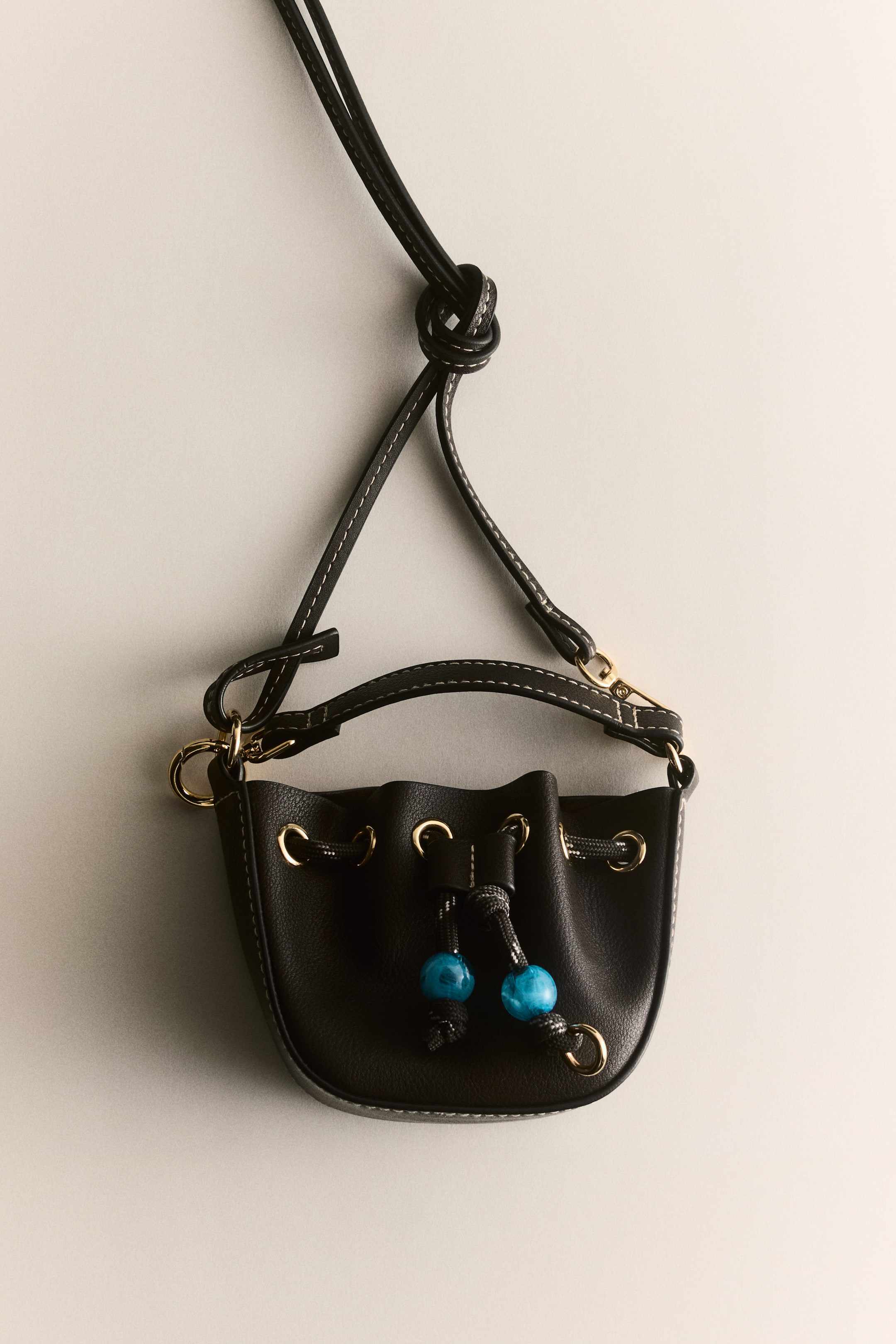 View larger image: A small black textured bucket bag is hanging, showcasing white contrasting stitching and gold-toned hardware. The drawstring closure is gathered at the top, featuring two striking turquoise spherical beads on a dark cord, with a sleek black strap attaching to the bag.
