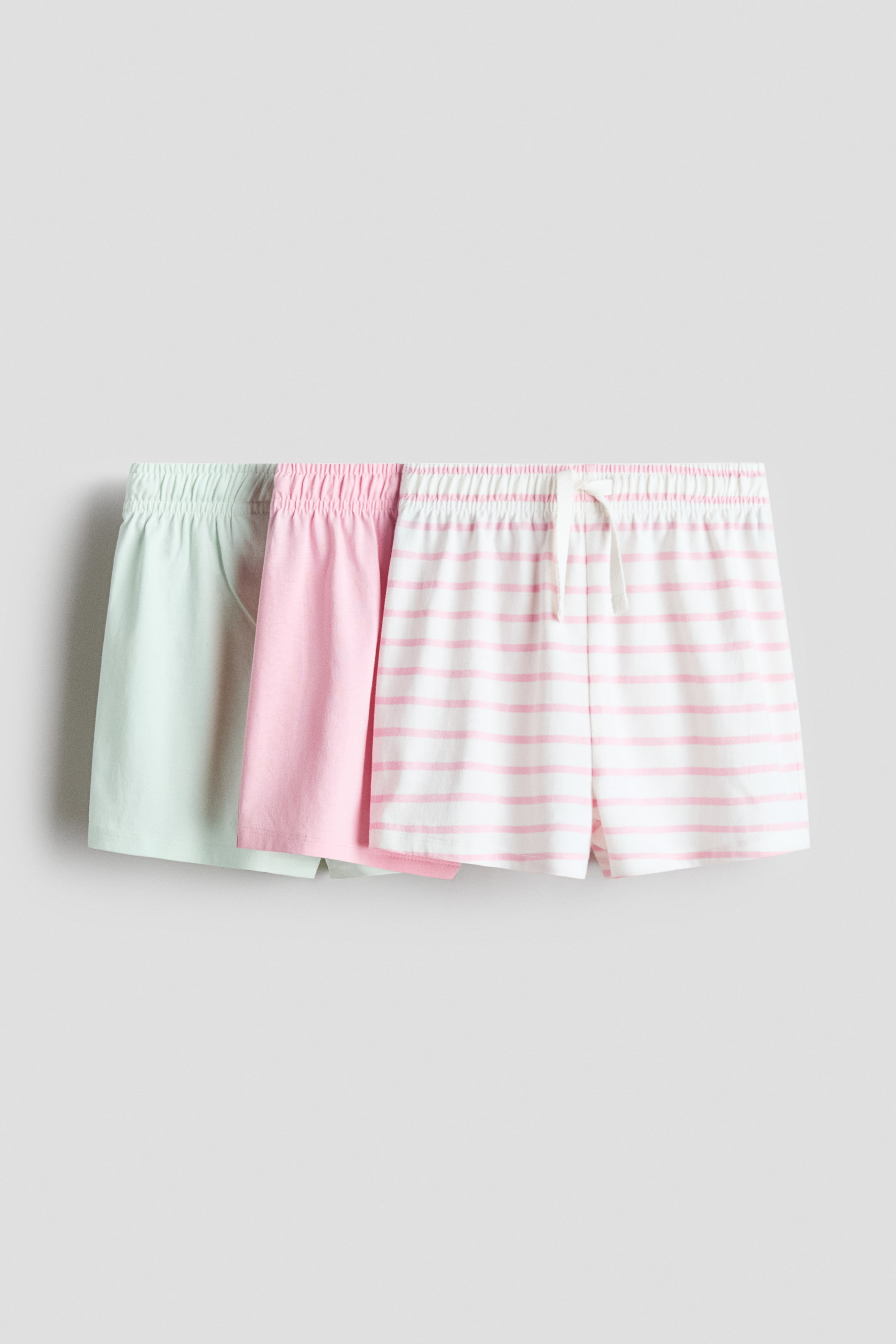View larger image: Three pairs of soft cotton jersey shorts are shown from the front, each with an elastic waistband. The pair on the left is light green, the pair in the middle is light pink, and the pair on the right is white with light pink horizontal stripes, featuring a white drawstring at the waist.