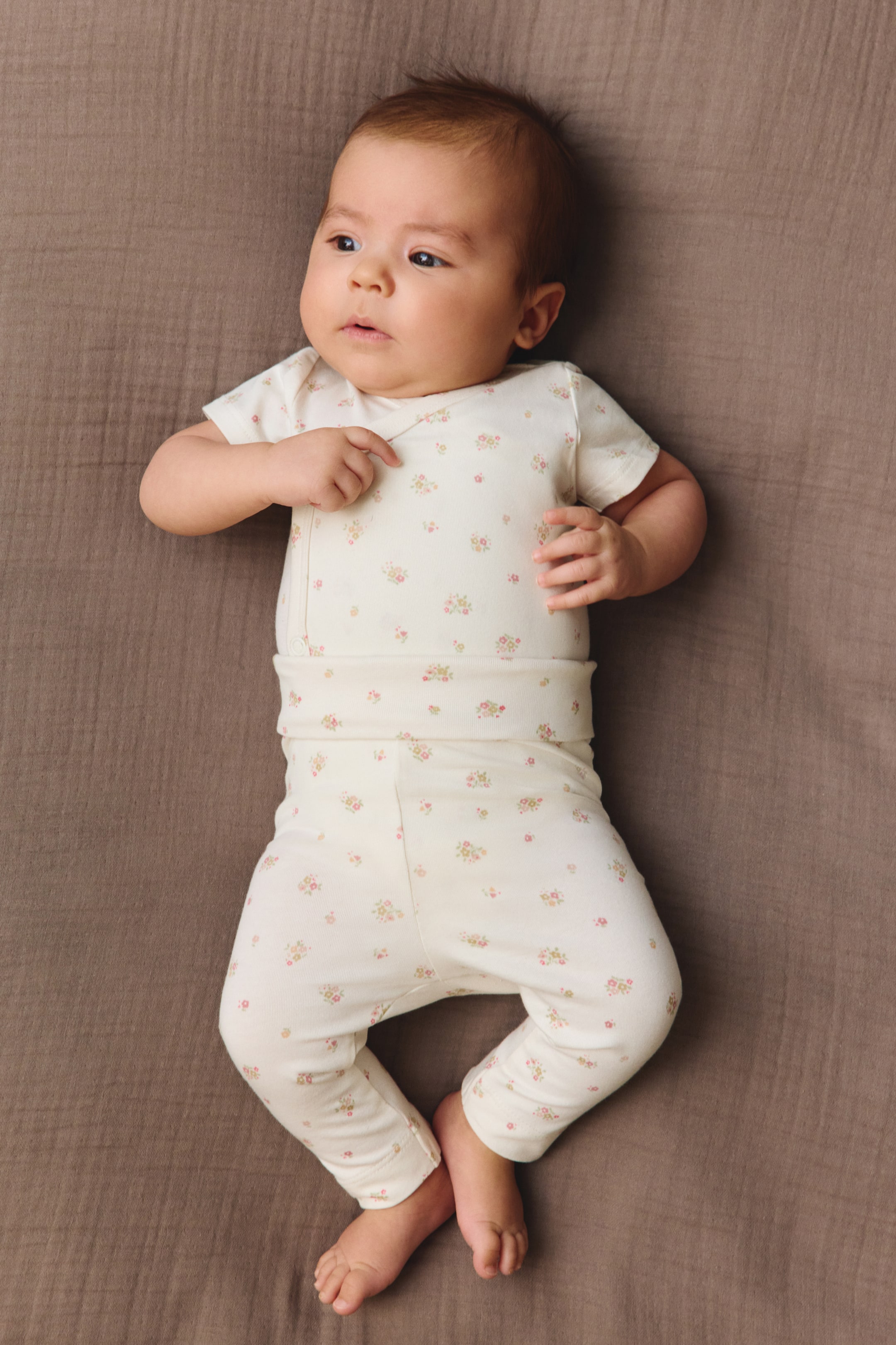 View larger image: A cream two-piece baby outfit is displayed from above, showcasing its front design and fit. It includes a short-sleeved top with a side snap closure and matching full-length pants with a wide, ribbed waistband, both adorned with a delicate pink and green floral print.