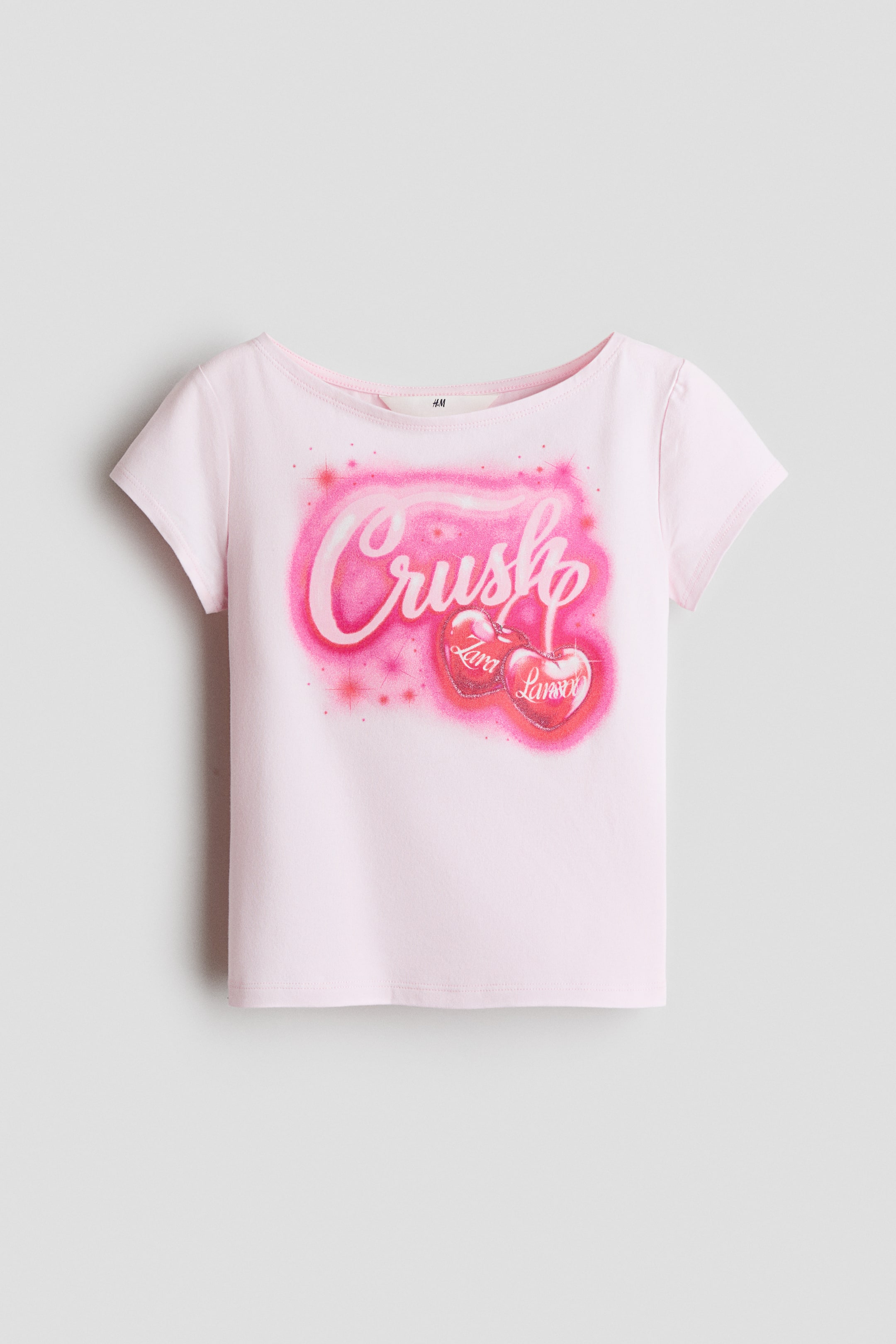 Voir une image plus grande: Le t-shirt rose pâle est présenté face avant, avec un col bateau et des manches courtes, arborant un graphique vibrant rose à l'aérographe sur le devant illustrant le mot "Crush" au-dessus de deux cerises en forme de cœur avec "Zara" et "Larsson" inscrits dessus.