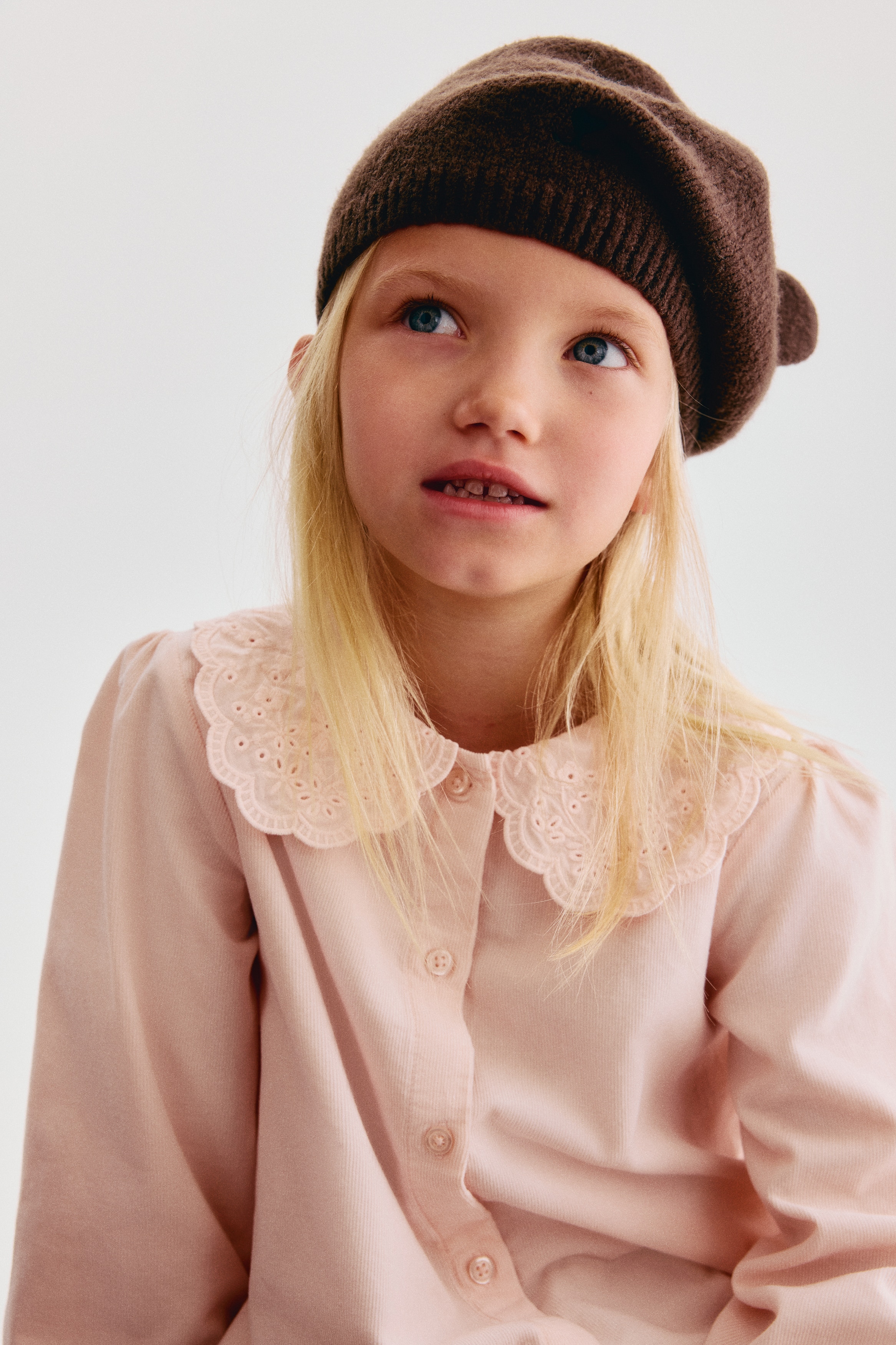 A dark brown knitted beret is shown from the front, featuring a circular, structured shape with two small, rounded animal ears attached near the top. The hat displays a soft, fine-knit texture with seams radiating from the centre crown.