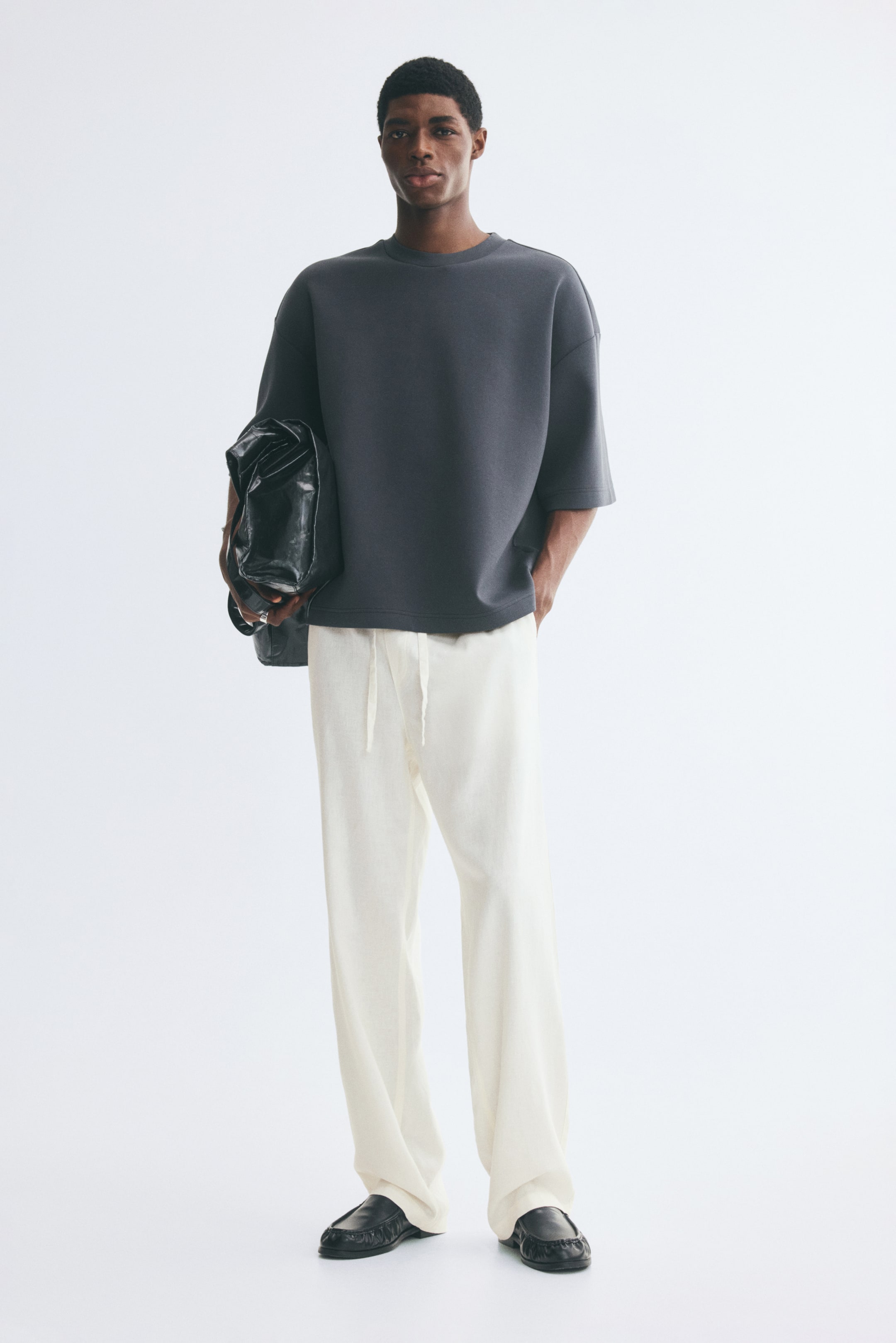 View larger image: Cream-coloured, relaxed-fit trousers made from a soft-weave fabric are worn by the model, featuring straight legs and an elasticated waist with a visible drawstring. The trousers are paired with a dark grey short-sleeved top, black slip-on shoes, and a black bag carried under the arm.