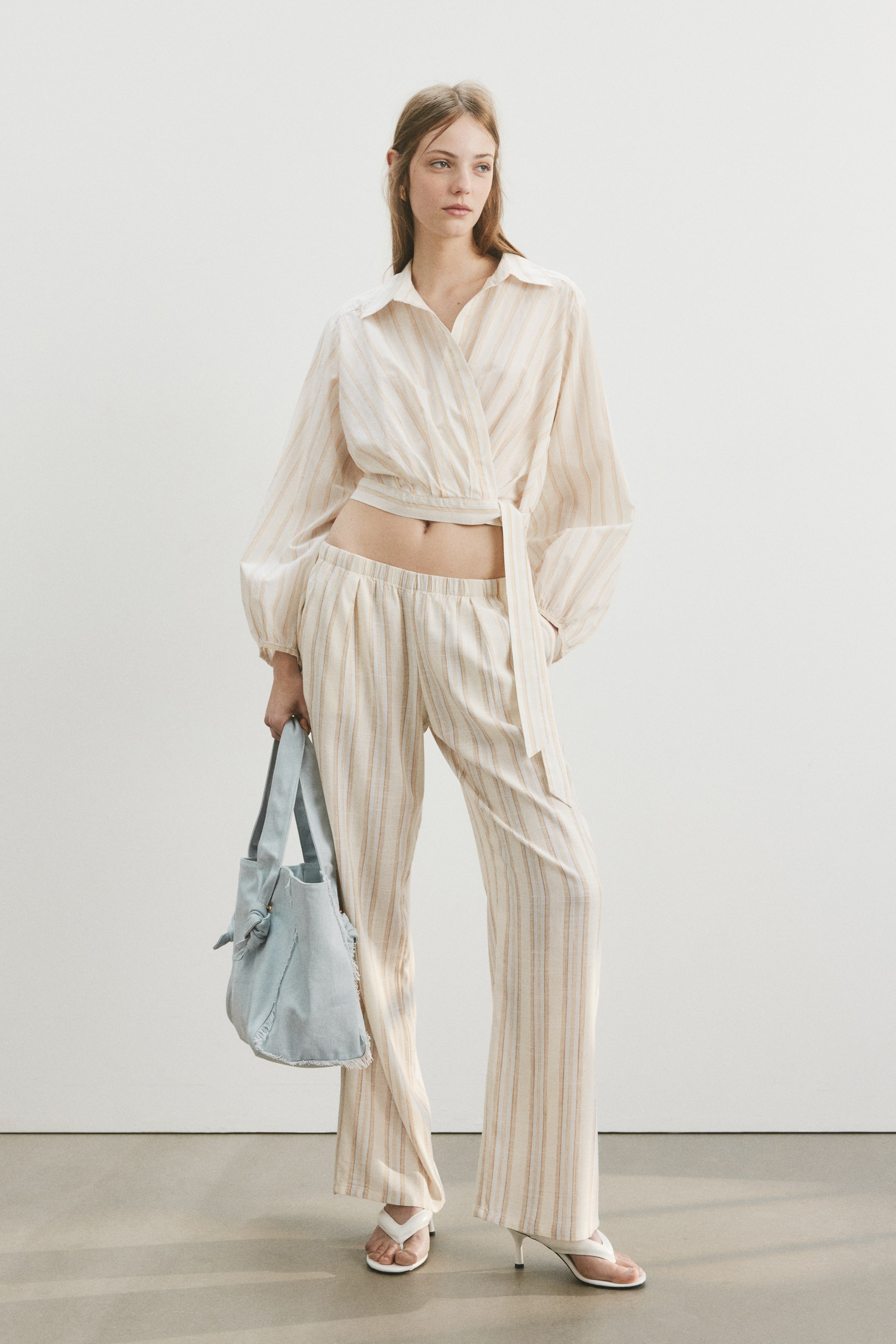 View larger image: Isang magkapares na set ng light beige at brown na striped na kasuotan ang ipinapakita, na binubuo ng isang wrap-front collared shirt na may mahabang manggas at tie closure, at wide-leg pants na may elasticated waistband. Ang cropped na shirt ay nagpapakita ng midriff. Isang light blue na malambot na tote bag na may knotted handles at puting high-heeled thong sandals ay kasama rin sa ipinapakita.