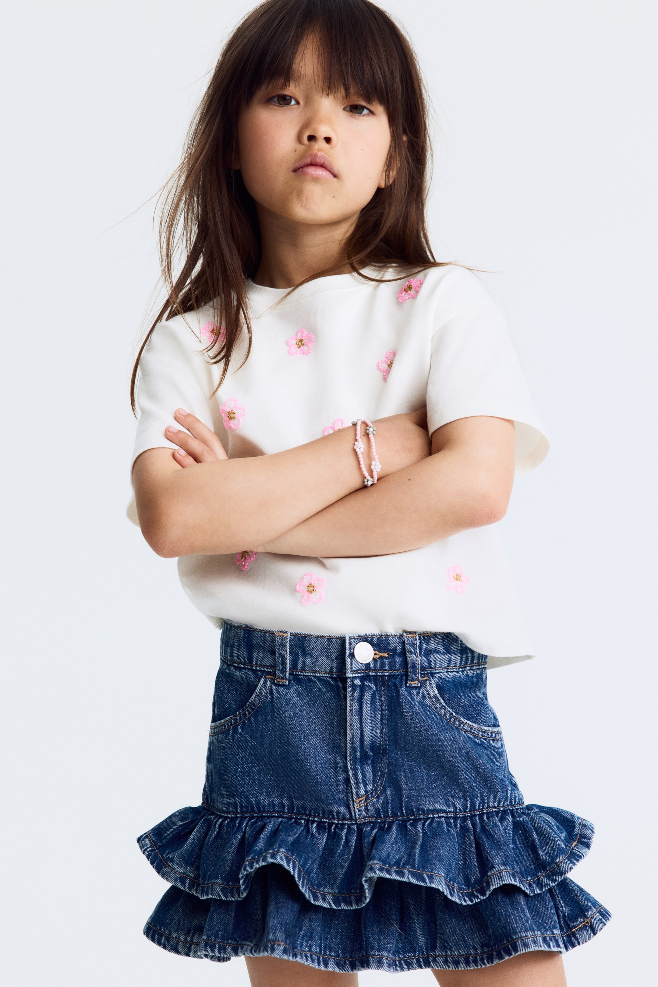Agrandir l'image: Un haut blanc à col rond et manches courtes présente des appliqués de fleurs roses brodées éparpillées. Il est porté avec une mini-jupe en denim bleu foncé, dotée d'un bouton visible à l'avant, de poches avant et de deux volants à l'ourlet.