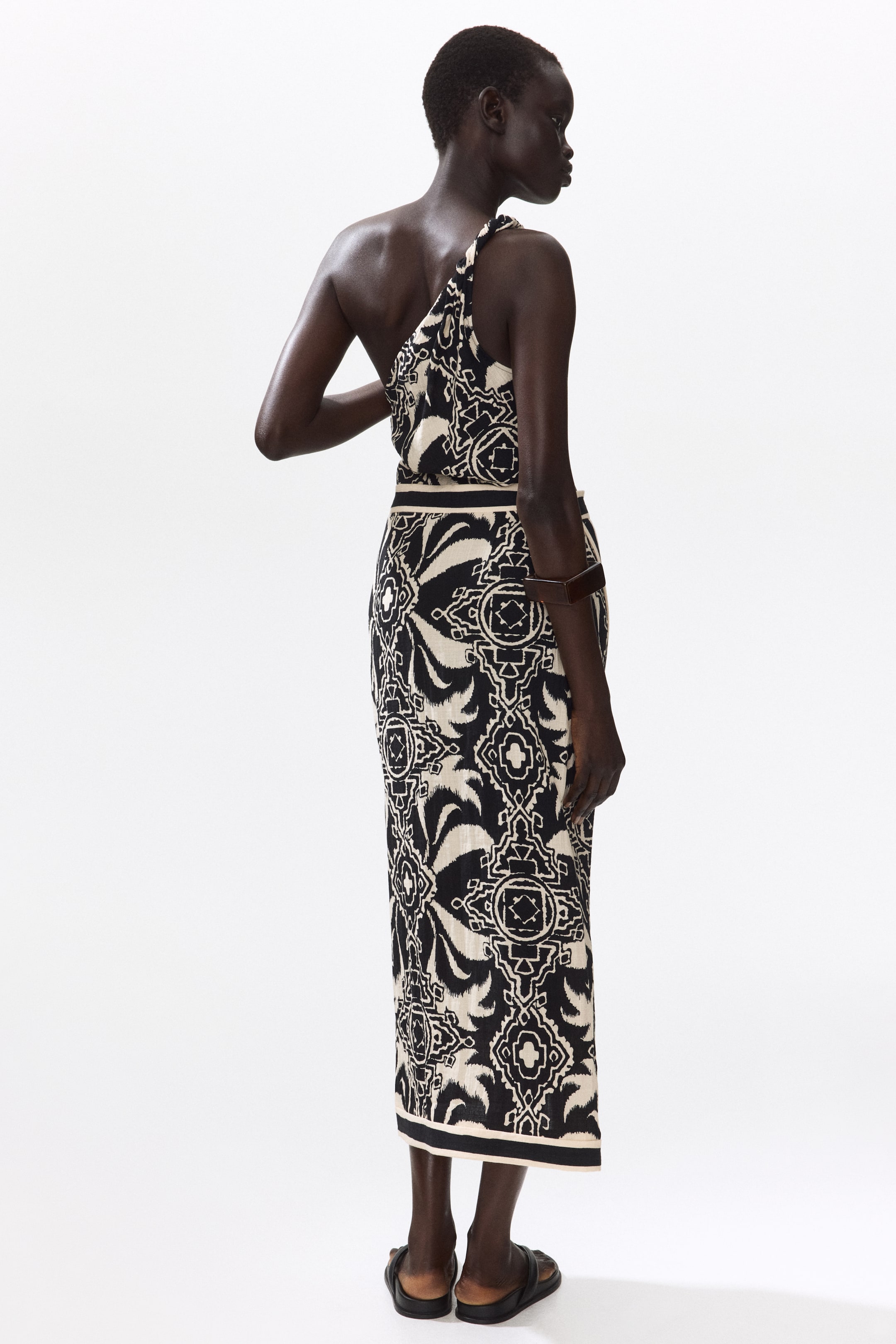 Größeres Bild ansehen: Ein One-Shoulder-Midikleid zeigt ein schwarz-weißes geometrisches und organisches Muster, das aus einem Blickwinkel von hinten rechts zu sehen ist. Das Kleid verfügt über einen verdrehten One-Shoulder-Träger, eine definierte Taille mit einem horizontalen Band und einen gesäumten Saum.