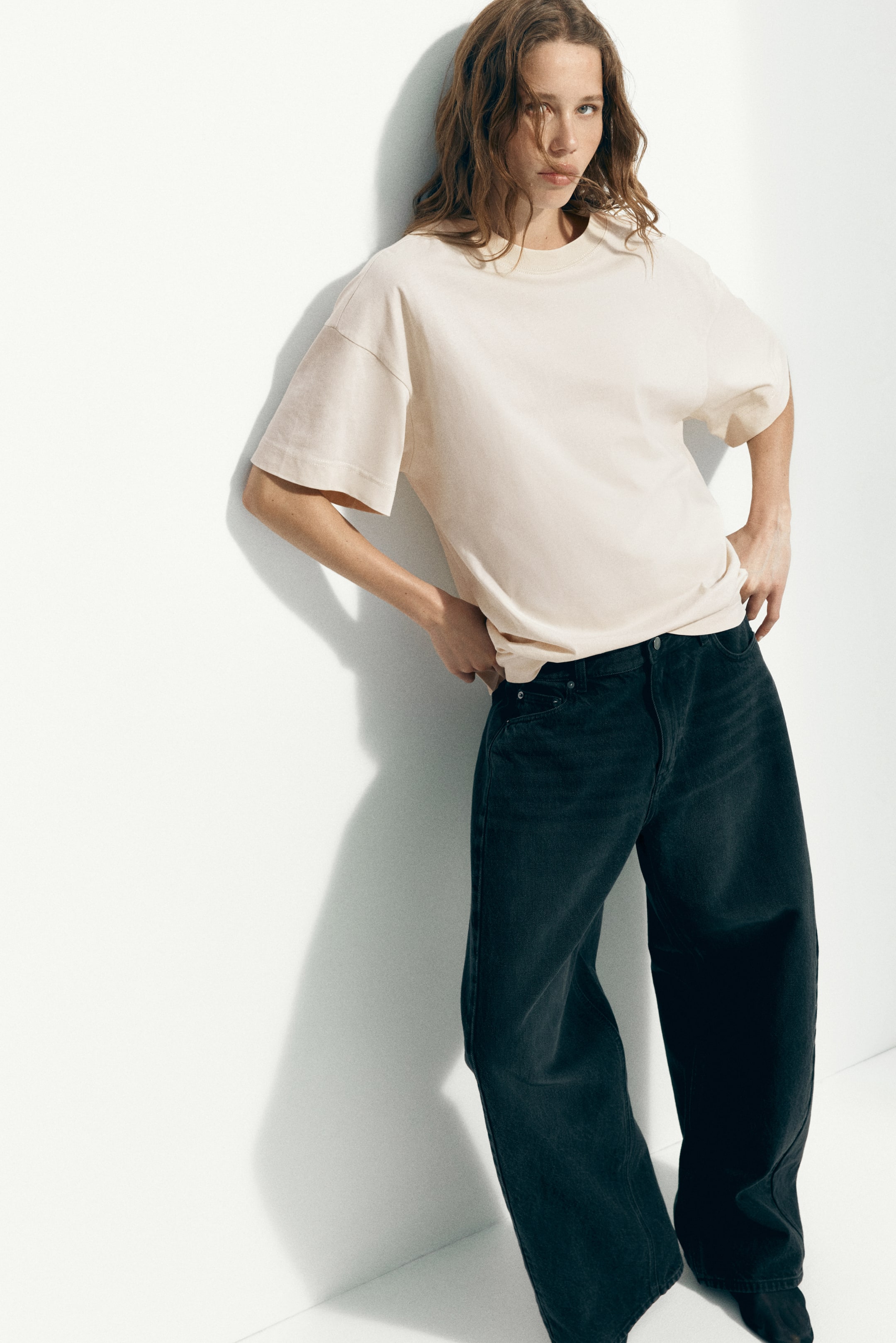 Grotere afbeelding bekijken: Een crèmekleurig, oversized T-shirt met ronde hals, verlaagde schouders en brede korte mouwen wordt gedeeltelijk ingestopt in donkergrijze, high-waisted, wijde jeans, wat een ontspannen pasvorm presenteert.