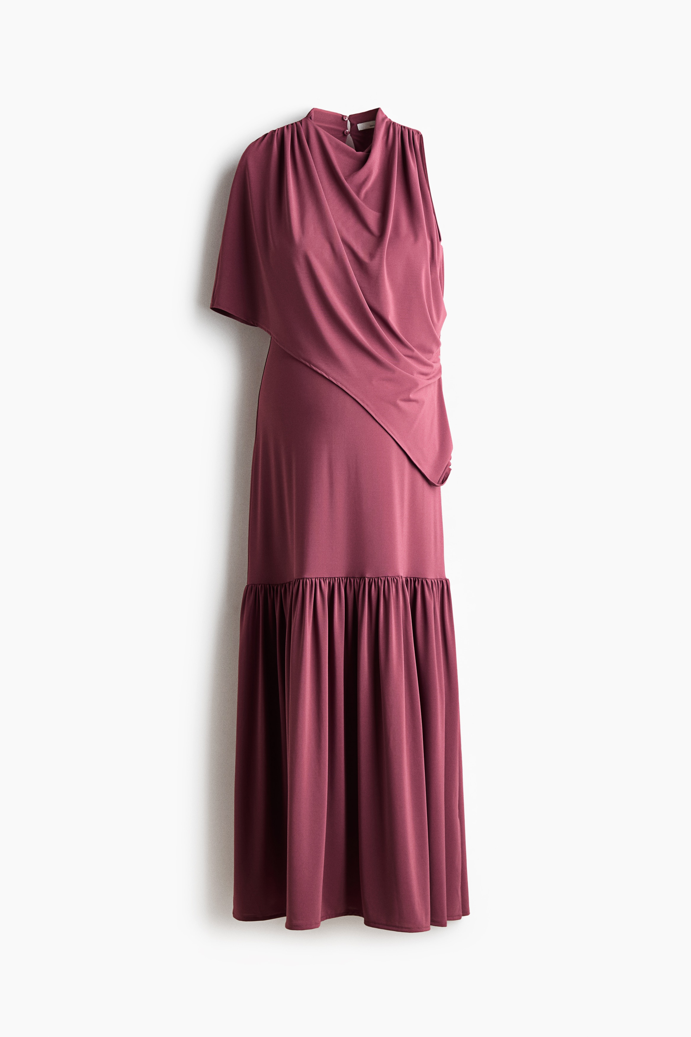 A long, sleeveless plum-coloured dress is being worn by the model, shown from a three-quarter back angle. The dress features an asymmetric cape-like layer draped over one shoulder, a keyhole opening at the back of the neck, and a gathered tier below the hip that flows into a wide, flared skirt. The model is styling the dress with light pink, floral-embellished high-heeled mules.