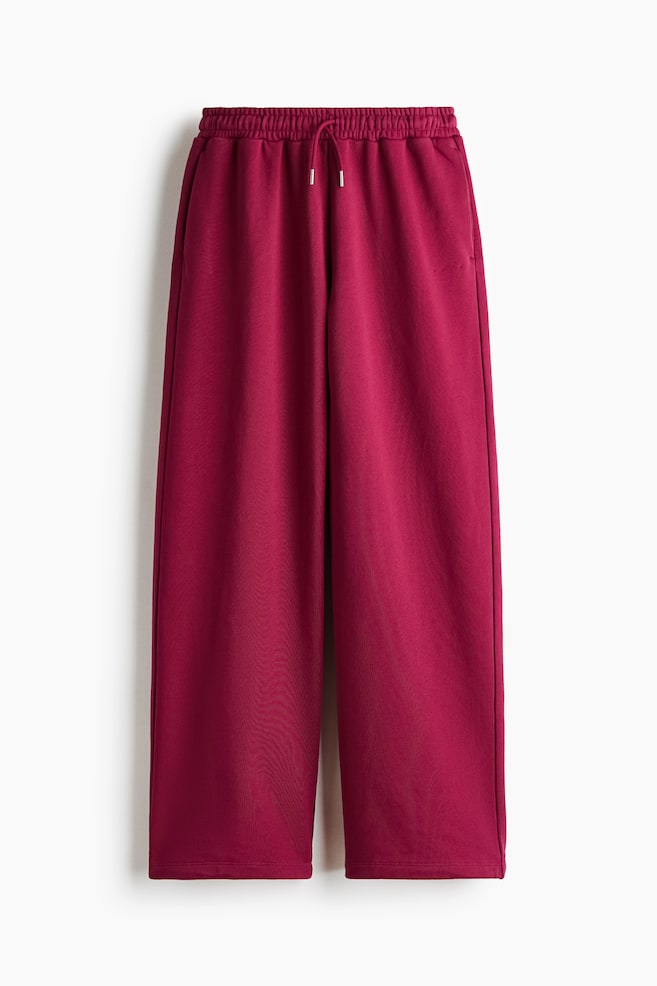 Ein Paar magentafarbene Sweatpants mit weitem Bein hÃ¤ngt frontal und zeigt einen gerafften elastischen Bund mit einem verstellbaren, farblich abgestimmten Kordelzug und silberfarbenen Metallenden. Die Hose fÃ¤llt in eine entspannte Wide-Leg-Silhouette mit sauberen SÃ¤umen und dezenten Seitentaschen in der Seitennaht. - 1