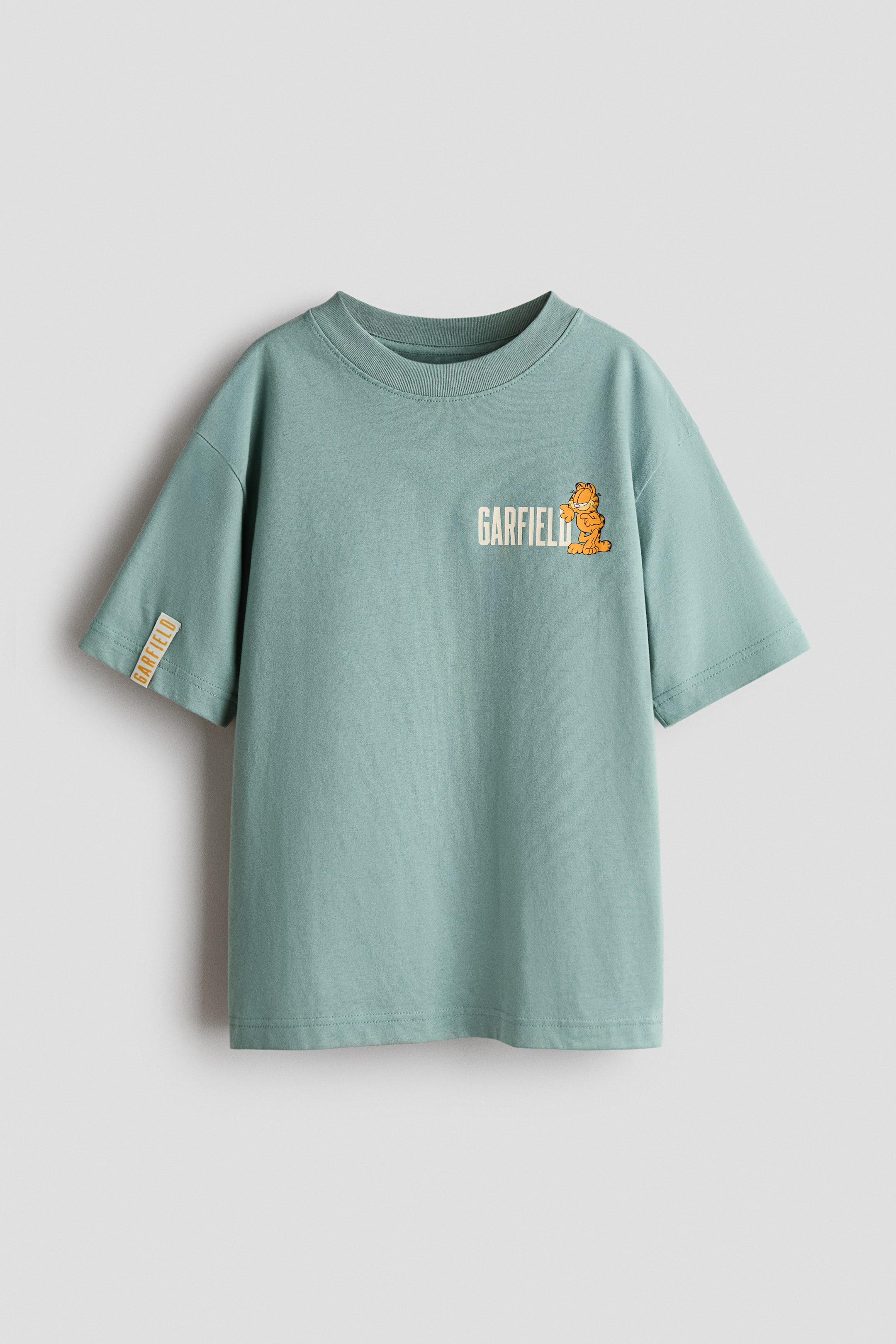 Xem ảnh lớn hơn: Một chiếc áo thun jersey cotton màu xanh xô thơm, chụp chính diện, đang hiển thị cổ tròn bo gân, vai trễ và tay ngắn. Áo có hình in đồ họa nhỏ nhân vật Garfield bên cạnh dòng chữ “GARFIELD” ở ngực trái, cùng một thẻ trang trí nhỏ ở cổ tay áo bên phải.