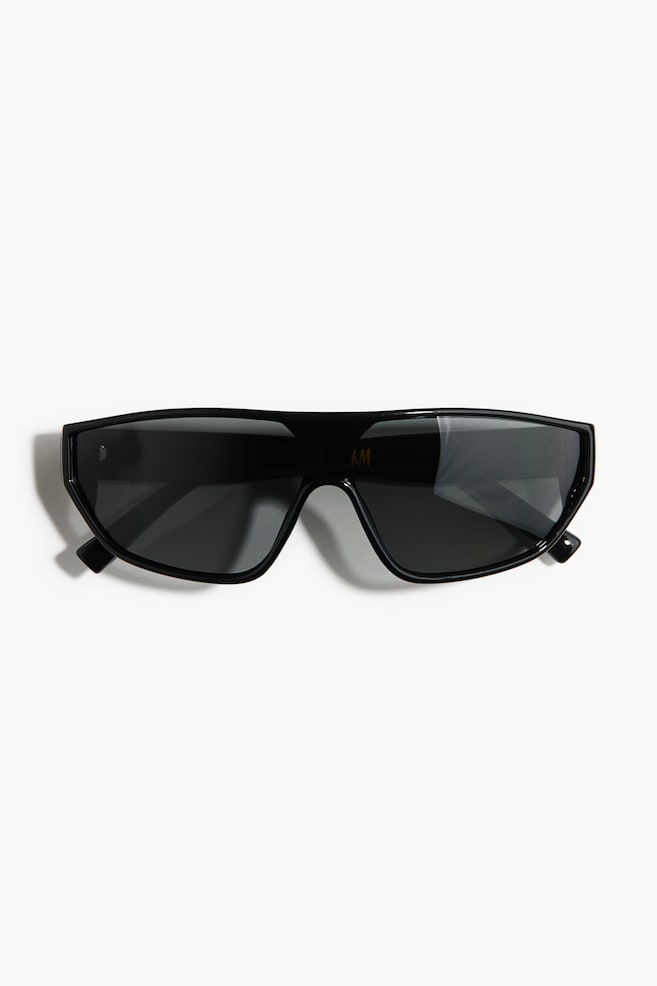 Des lunettes de soleil style bouclier noires sont posées à plat, présentant une monture épaisse et brillante avec des verres foncés. Un petit logo 'H&M' est visible sur le verre droit, et un détail métallique circulaire est visible sur la branche gauche. - 1