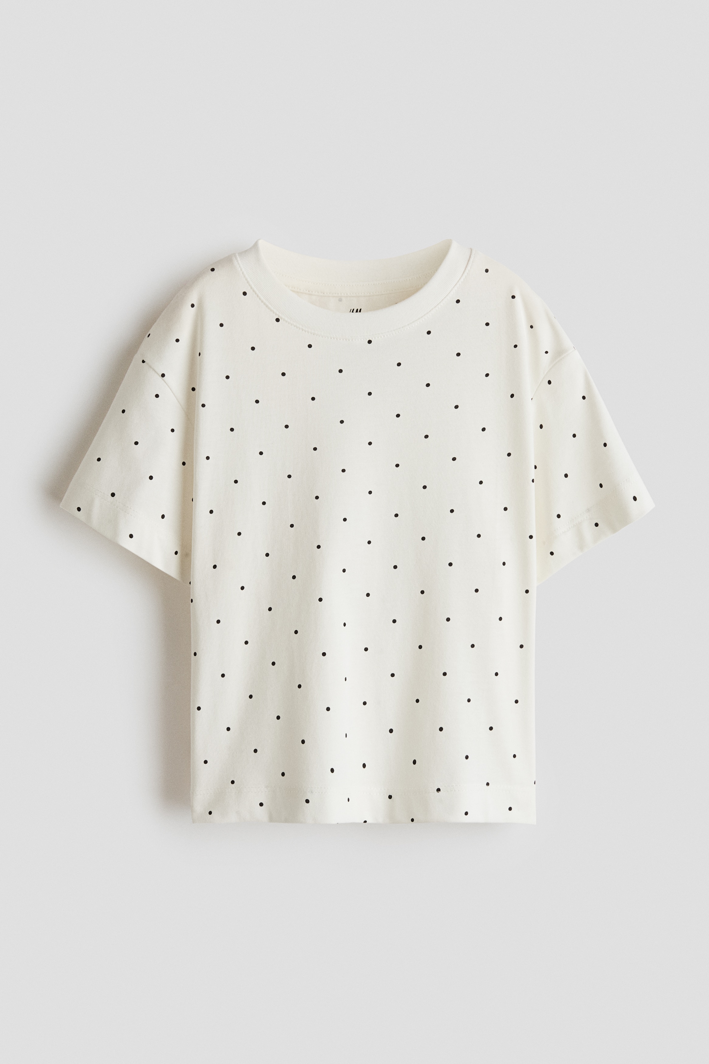 An off-white, short-sleeved t-shirt featuring a pattern of small black polka dots is being worn by the model. The t-shirt has a crew neckline and a relaxed fit with dropped shoulders, and is styled with black pants.