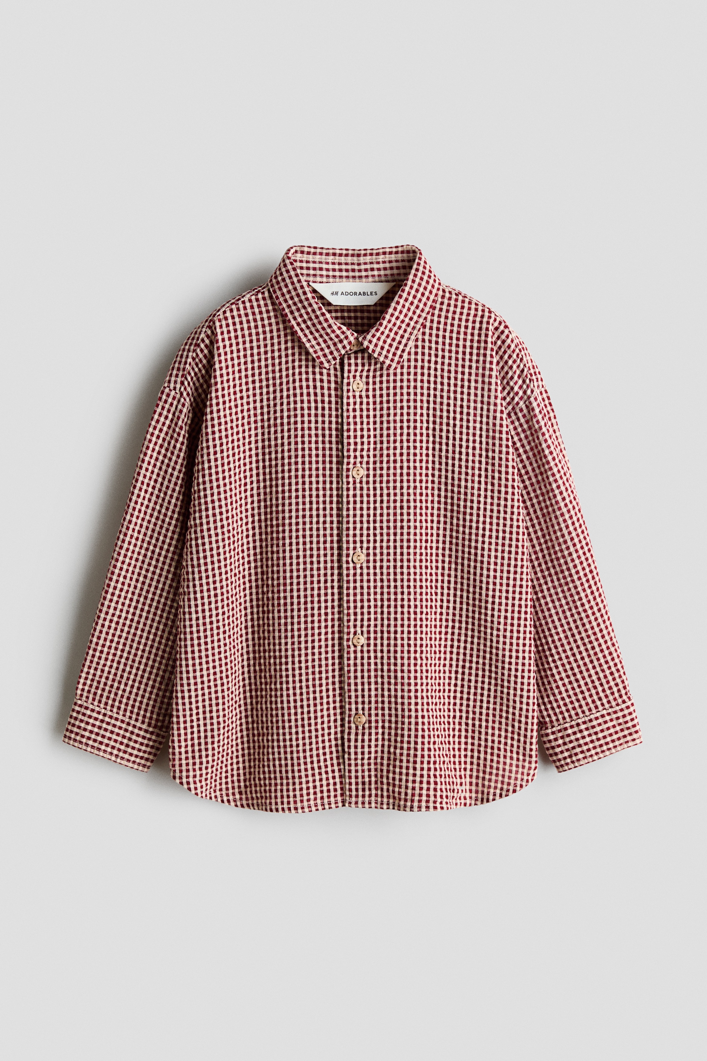 H&M Adorables koleksiyonundan uzun kollu bir gömlek, dokulu müslin kumaŠüzerinde kırmızı ve beyaz gingham desenle önden gösterilmektedir. Gömlek, beyaz bir tiÅörtün üzerine kısmen düÄmeleri açık Åekilde giyilerek, klasik yakası ve kahverengi düÄme kapamaları vurgulanmaktadır.