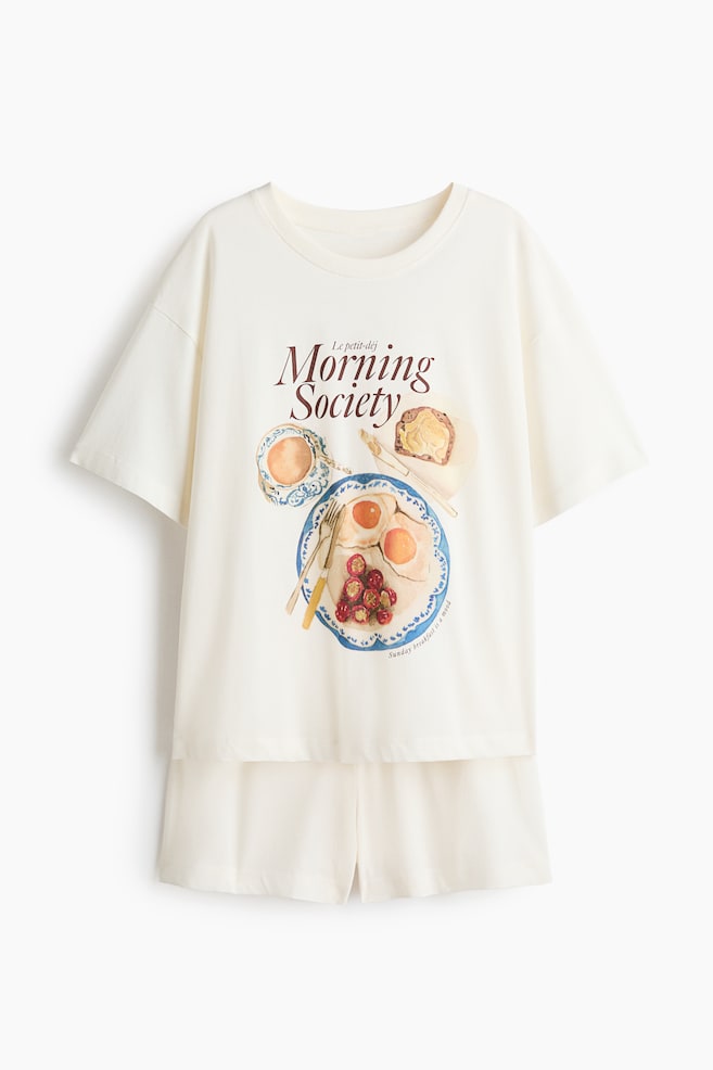 Pyjamas comprising a T-shirt and shorts are displayed from the front. The cream T-shirt features a ribbed trim around the neckline, dropped shoulders, and a breakfast-themed graphic print, including the text "Le petit-déj Morning Society", a cup, toast, and a plate with fried eggs and berries. The matching cream shorts display a covered elasticated waist. - 1