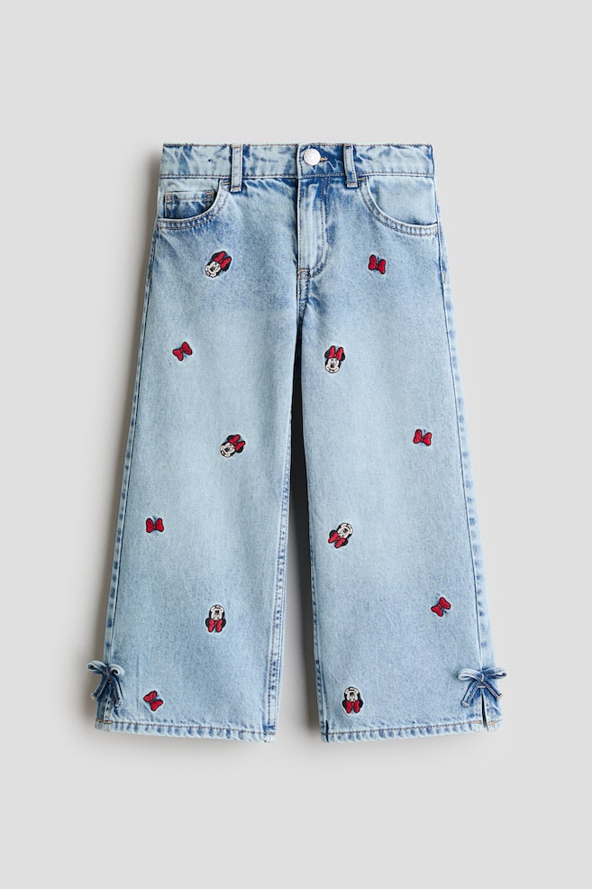 Light blue wide-leg denim jeans are laid flat, showcasing a front view with embroidered Minnie Mouse heads and red bows scattered across both legs. Small denim bows tie the side slits at the leg openings. - 1