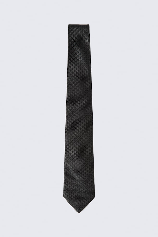 A Jacquard Weave Silk Tie in dark grey is hanging vertically, displaying a subtle, repeating geometric jacquard pattern across its entire length. The tie is widening slightly toward the bottom and ending in a classic pointed tip. - 1