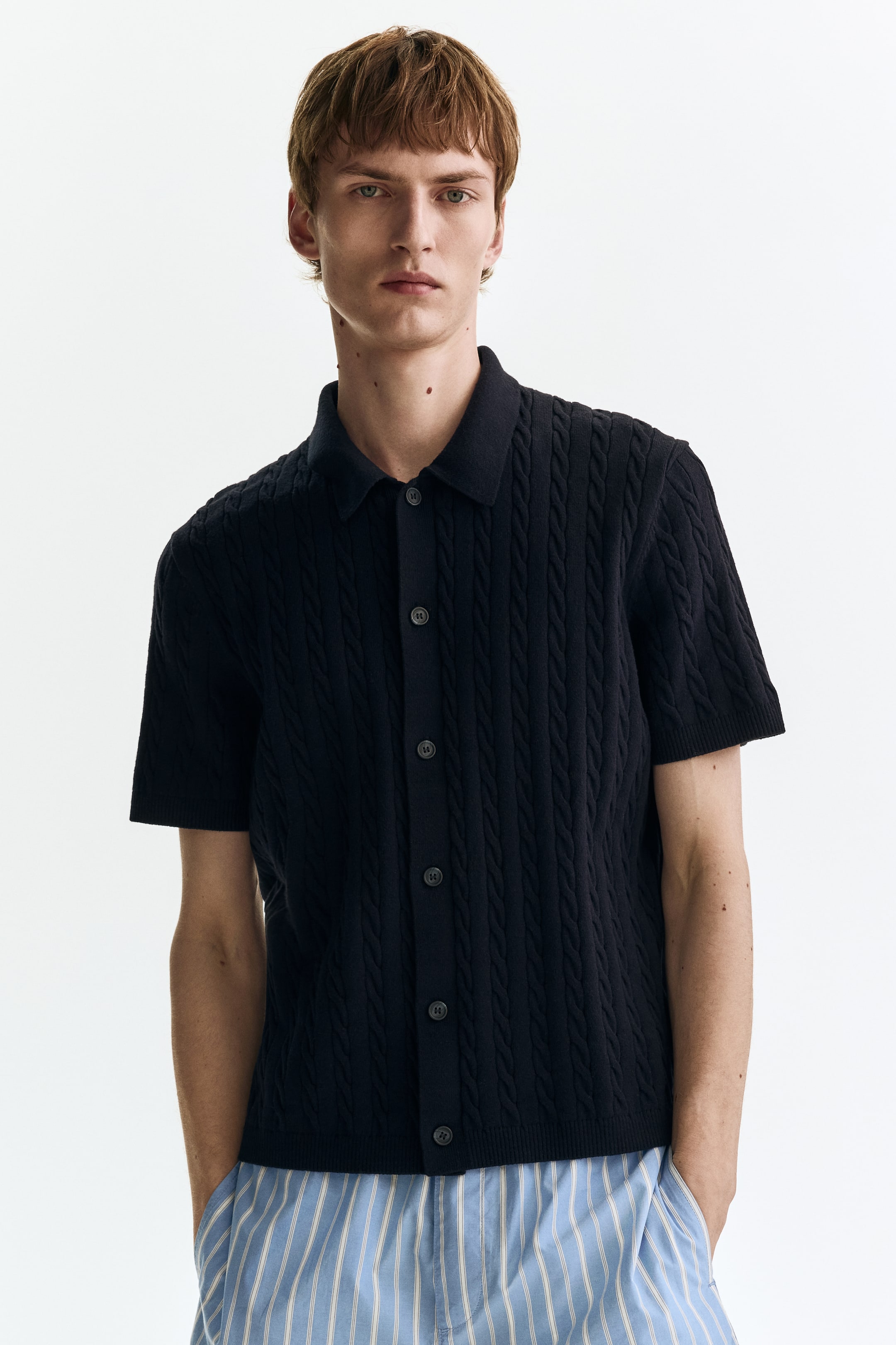 Ver imagen más grande: Se presenta una camiseta polo de punto de manga corta en color negro desde el frente, con un diseño de punto trenzado en toda la prenda, un clásico cuello de polo y una tapeta de botones completa con botones oscuros.