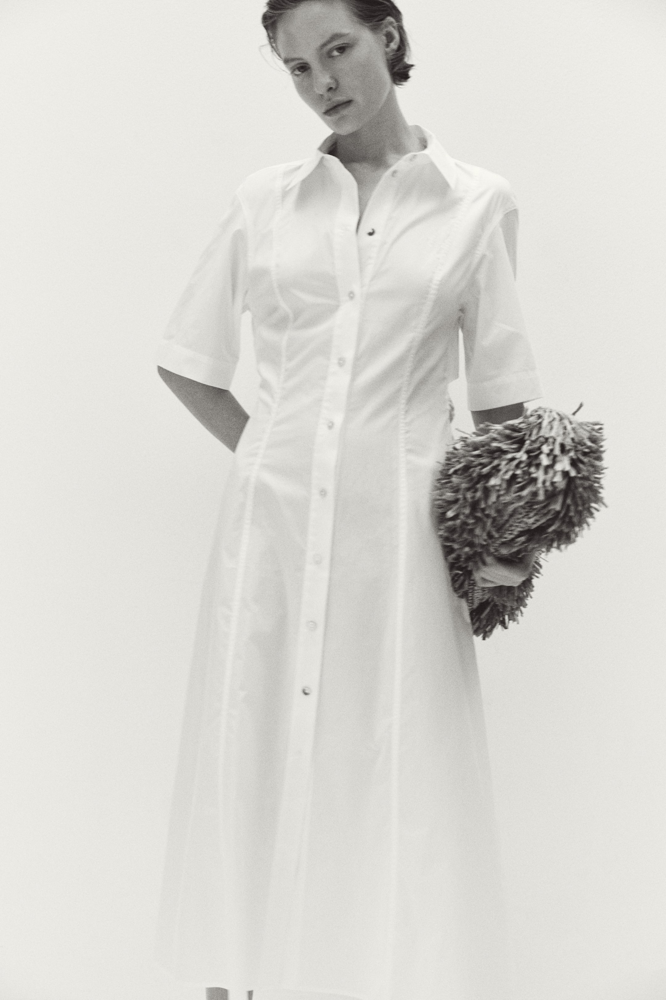 View larger image: The model is wearing a calf-length dress that showcases a collar, buttons running down the front, subtly dropped shoulders, elbow-length sleeves, and noticeable seams along the front. The skirt has a gentle flare, and the model is holding a textured item in their right hand.